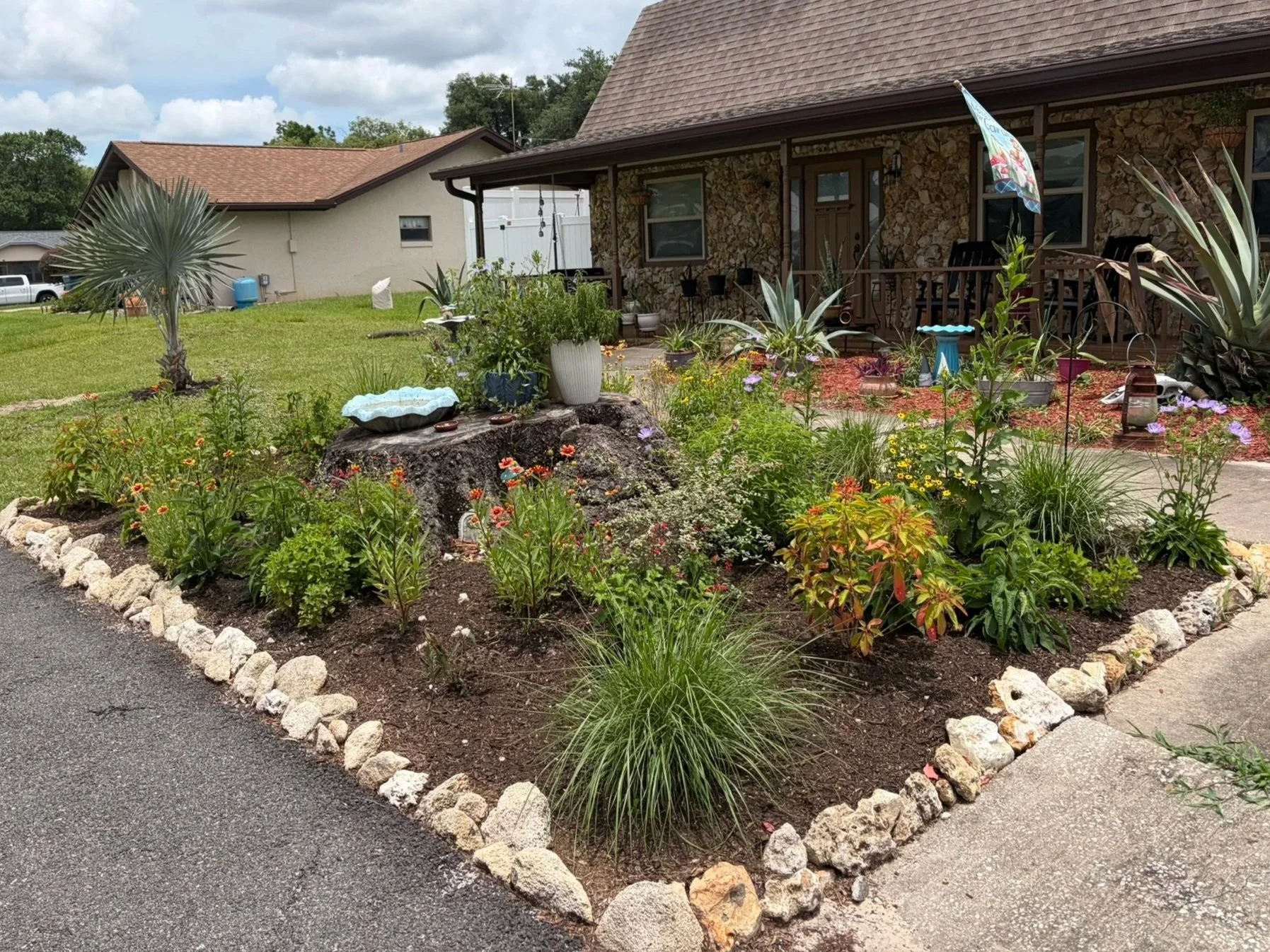 A backyard garden with various plants and flowers, bordered by large rocks, in front of a house with a stone porch and brown roof.