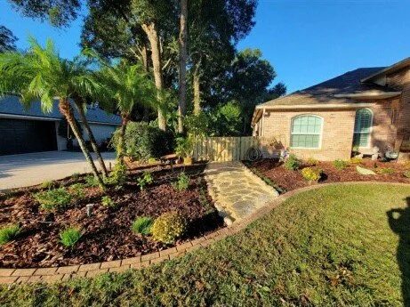 A landscaped front yard with a stone walkway, lush green grass, and various plants and trees next to a residential house under a clear blue sky.