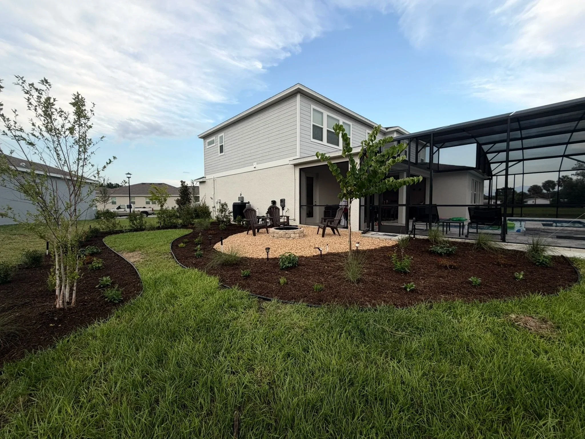 A suburban backyard with a landscaped garden, green grass, small trees, and bushes, a patio with chairs around a fire pit, a screened-in pool area, and a two-story house in the background with a blue sky and some clouds overhead.