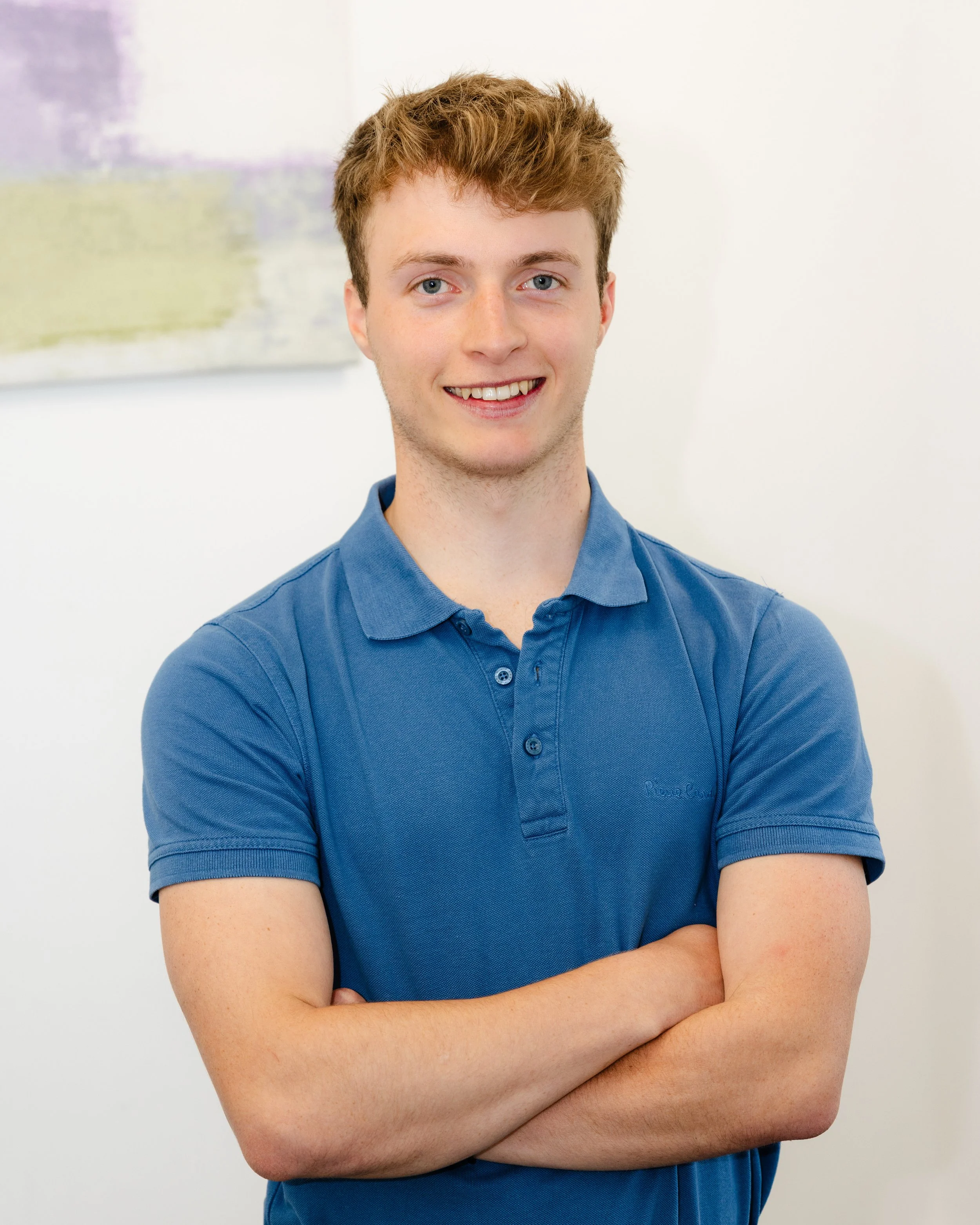 A young man with light skin, short red hair, and blue eyes is smiling with arms crossed, wearing a blue polo shirt, standing in front of a white wall with a blurred abstract painting.