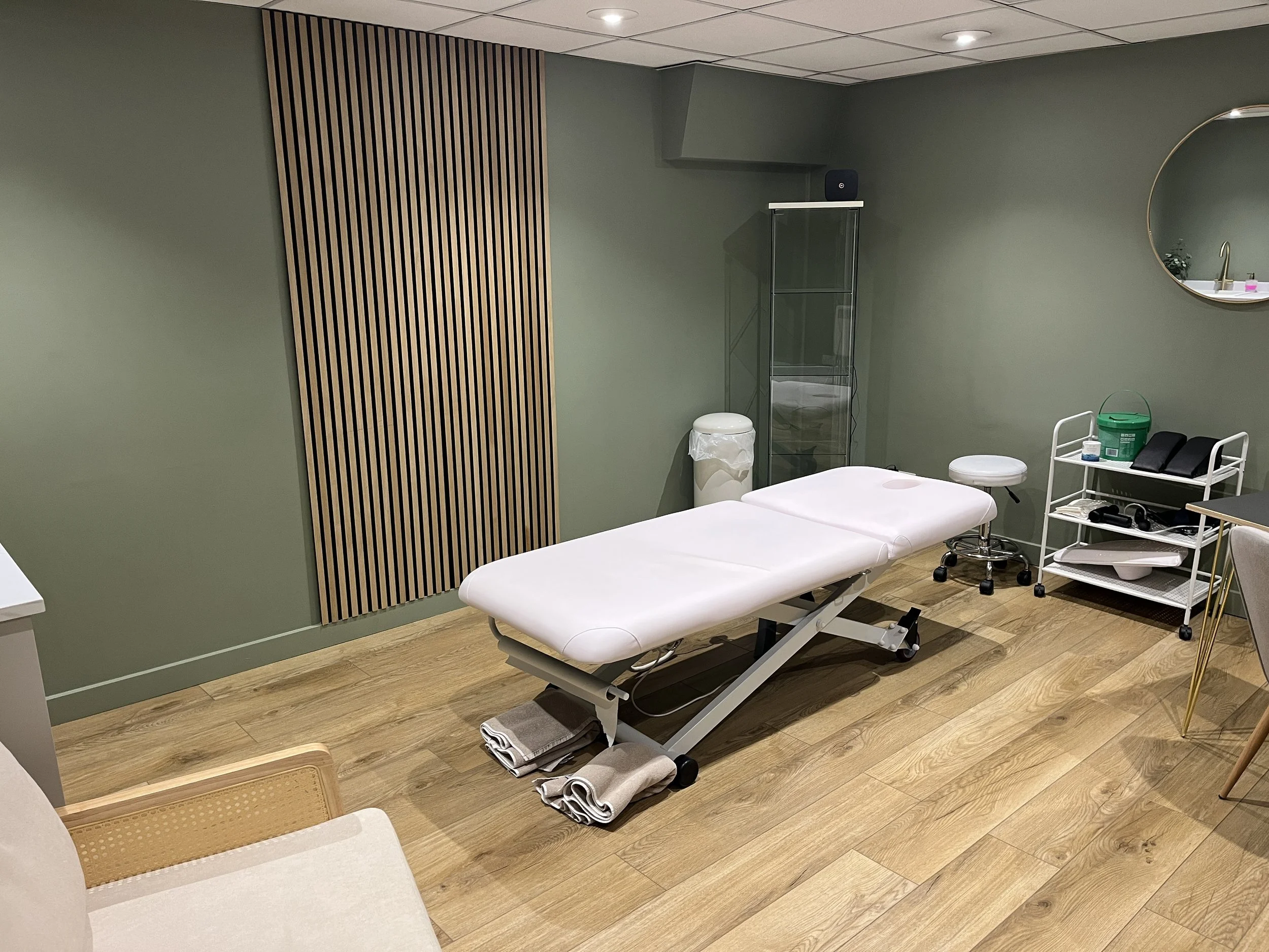 A treatment room with a massage or examination table, a rolling stool, storage cart with supplies, mirror, and decorative wooden wall panel.