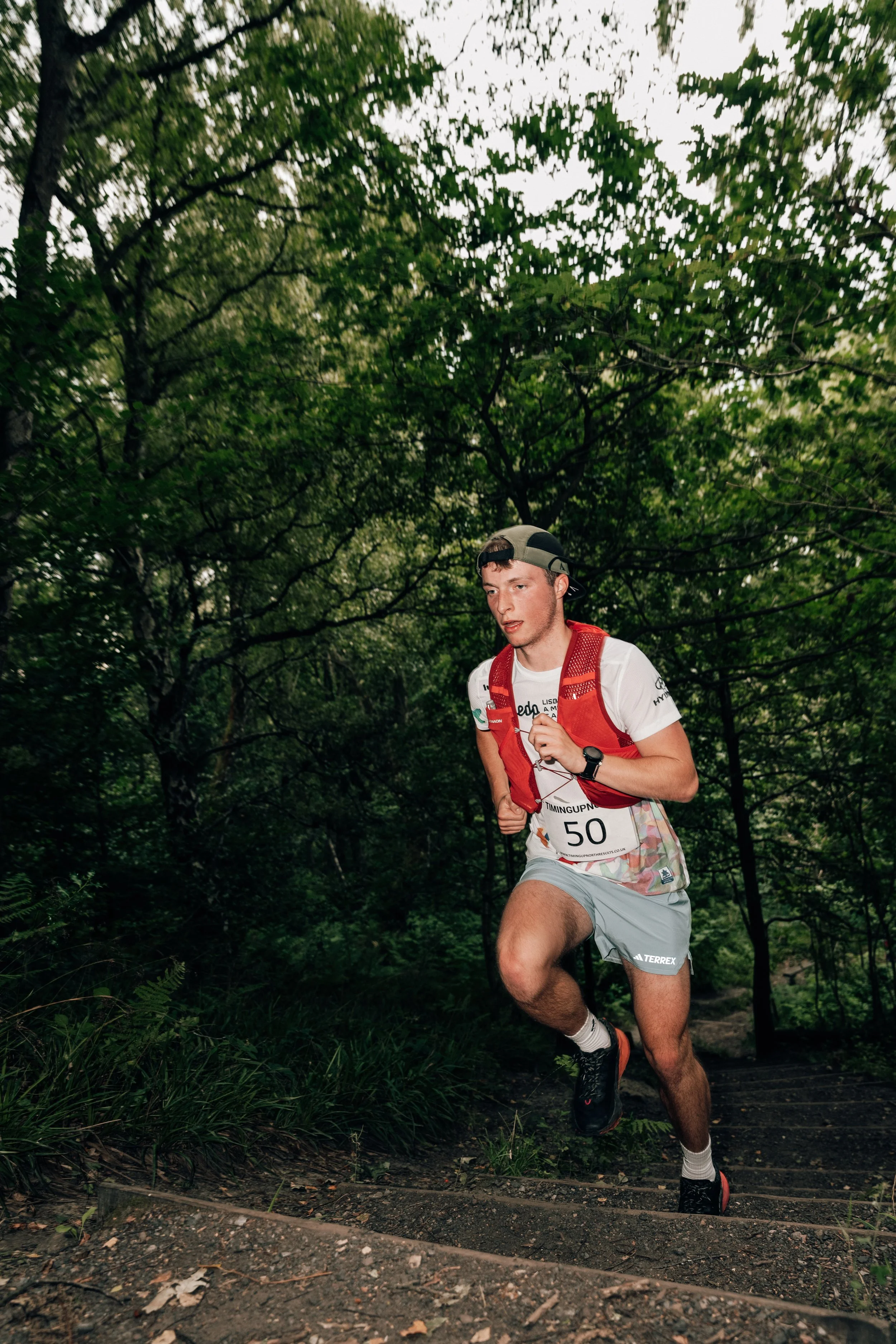 A male runner wearing a white shirt, gray shorts, black running shoes, a red hydration pack, and a black cap runs up a wooded trail.