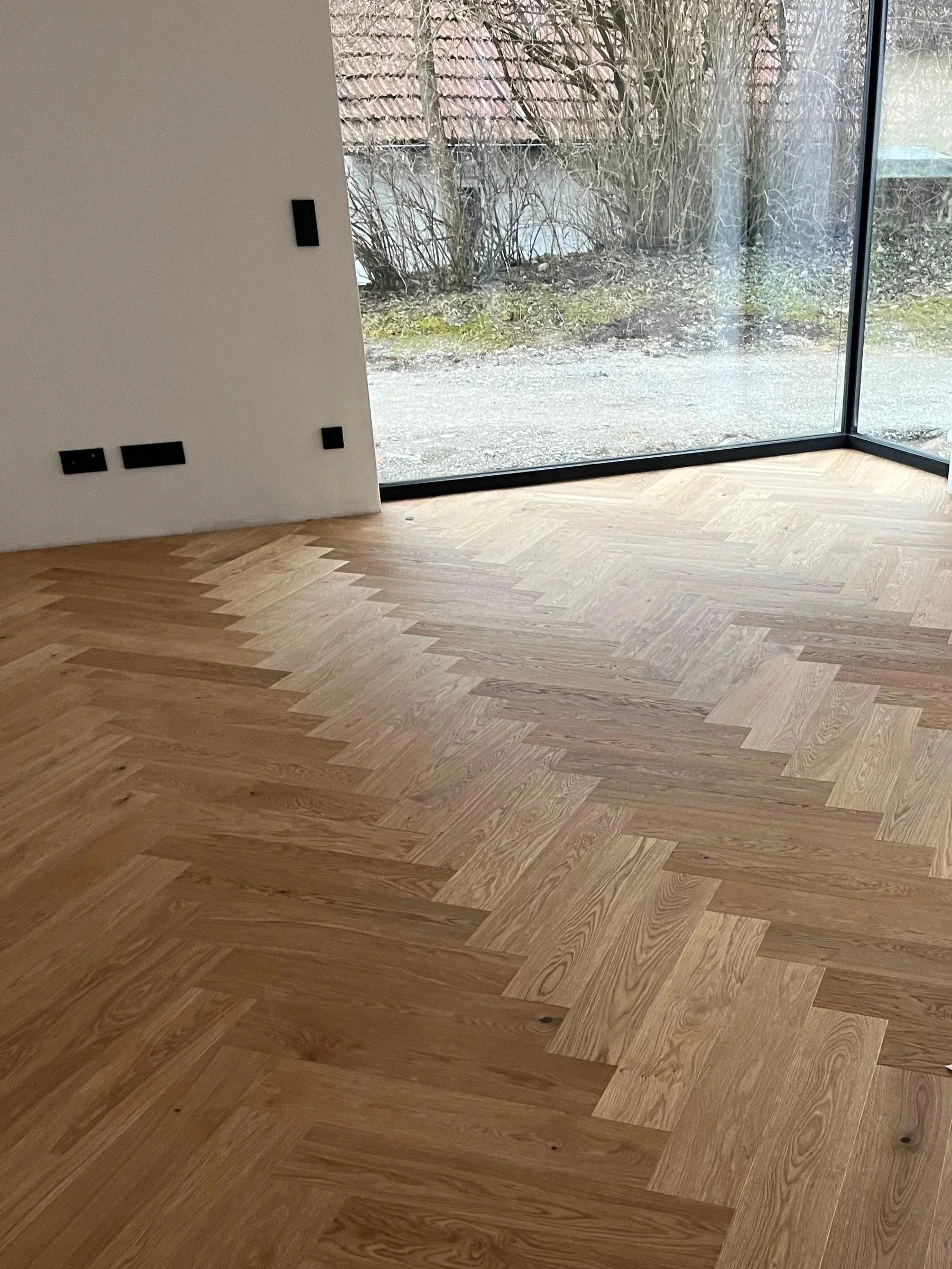 Innenansicht eines Raumes mit Holzboden und großem Fenster, durch das ein Garten mit Büschen und einem Weg zu sehen ist.