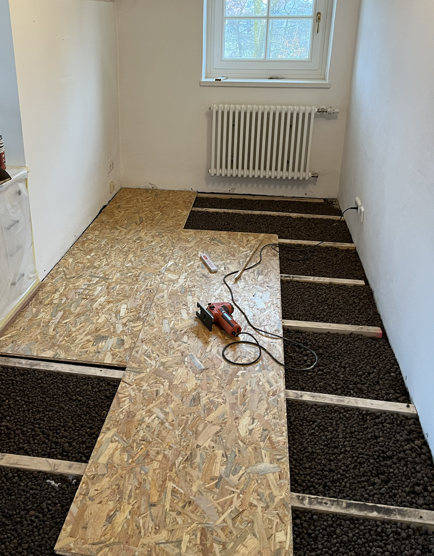 Ein Raum mit hölzernen Unterbodenplatten und Wärmeisolierung, während der Renovierung. Es liegen Baumaterialien und ein elektrisches Werkzeug auf dem Boden, mit einem Fenster und einem Heizkörper an der Wand.