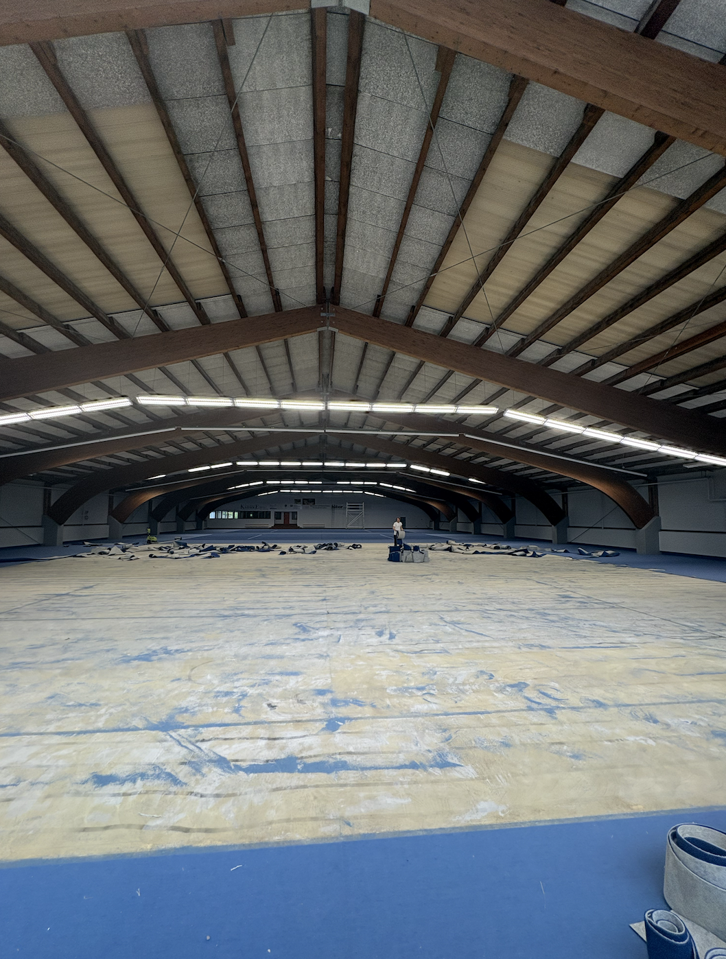Innenaufnahme einer großen Sporthalle, vermutlich Eisstadion, im Bau oder Renovierung befindlich, mit einer Person in der Mitte, die an der Eisfläche arbeitet oder sie vorbereitet.