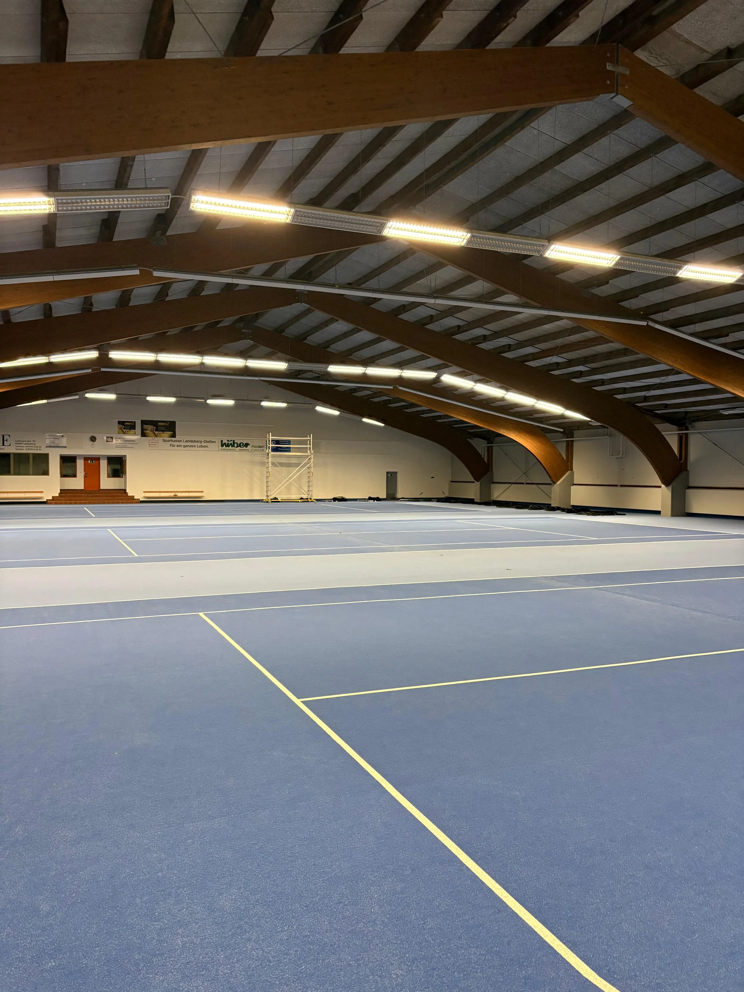 Leerer Sporthalle mit blauen Tennis-Courts, Holzdecke und Beleuchtung.
