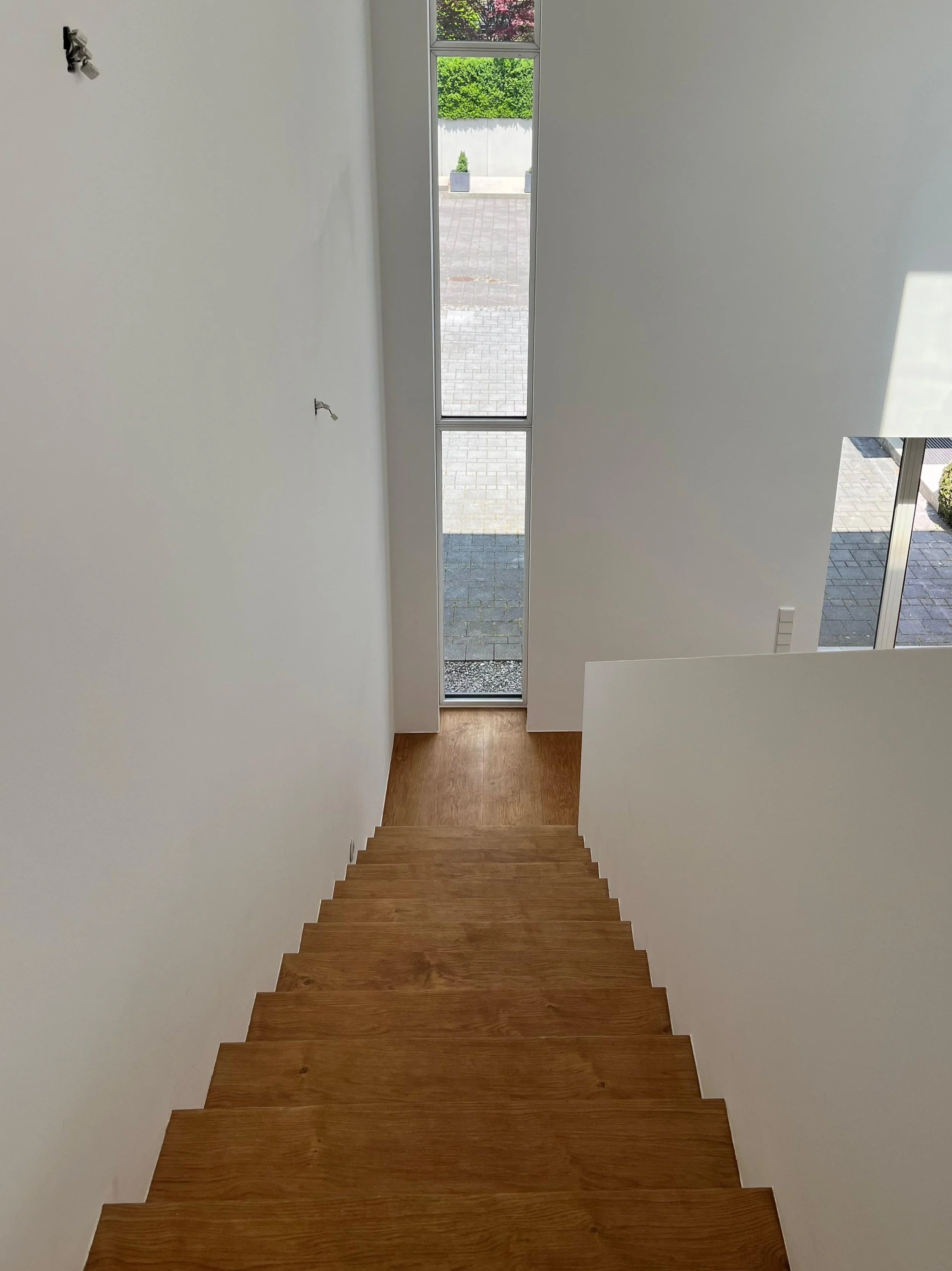 Innenansicht eines Hauses mit Treppe aus Holz, looking out through tall, narrow window at den Außenbereich mit gepflastertem Hof und grünen Büschen.