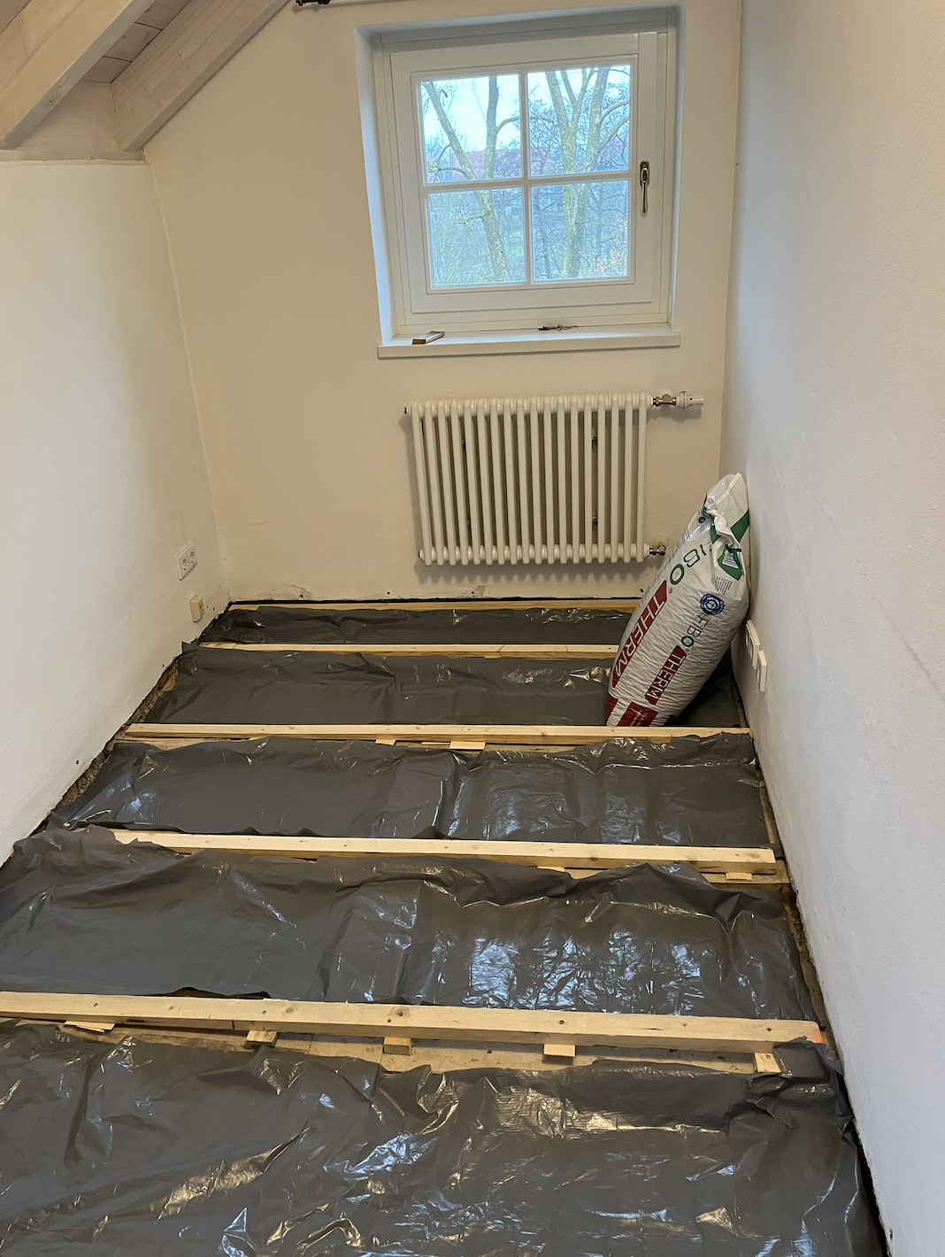 Ein leerer Raum mit einer Heizkörper unter einem Fenster, der Boden ist für eine Renovierung vorbereitet, mit schwarzen Abdeckfolien und Holzlatten auf dem Boden, eine Packung Dämmmaterial steht in der Ecke.