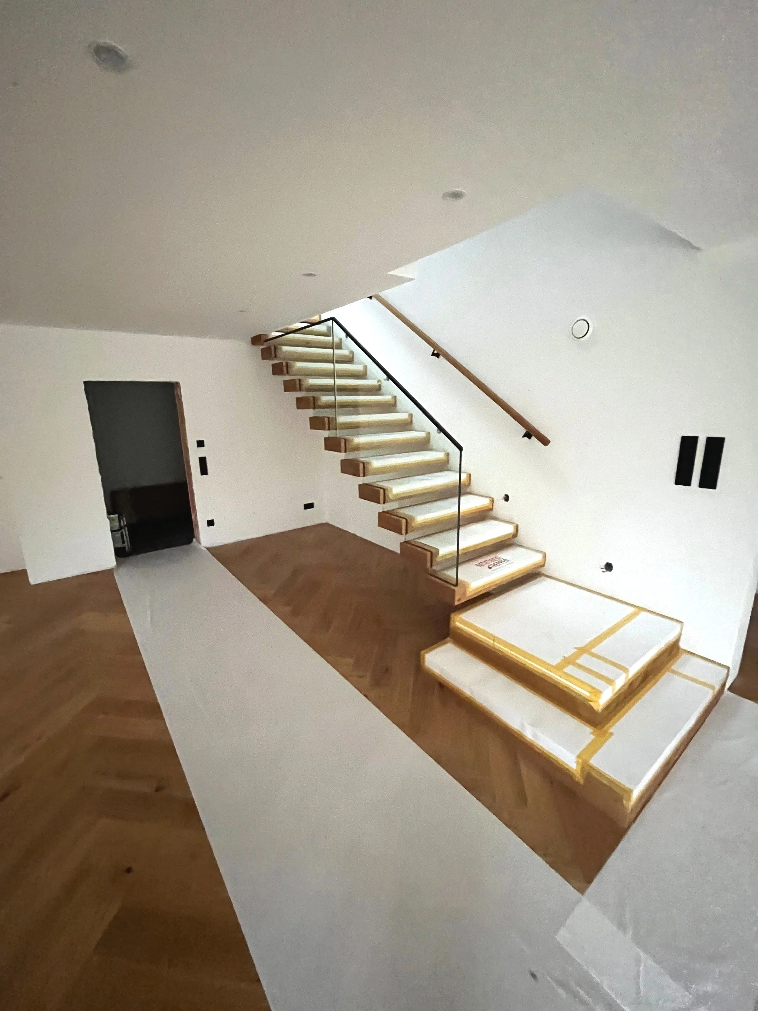 Innenansicht eines Hauses mit Holztreppe, die an einer weißen Wand ohne Geländer führt, in einem Raum mit Holzboden und mehreren Anschlussdosen an der Wand.