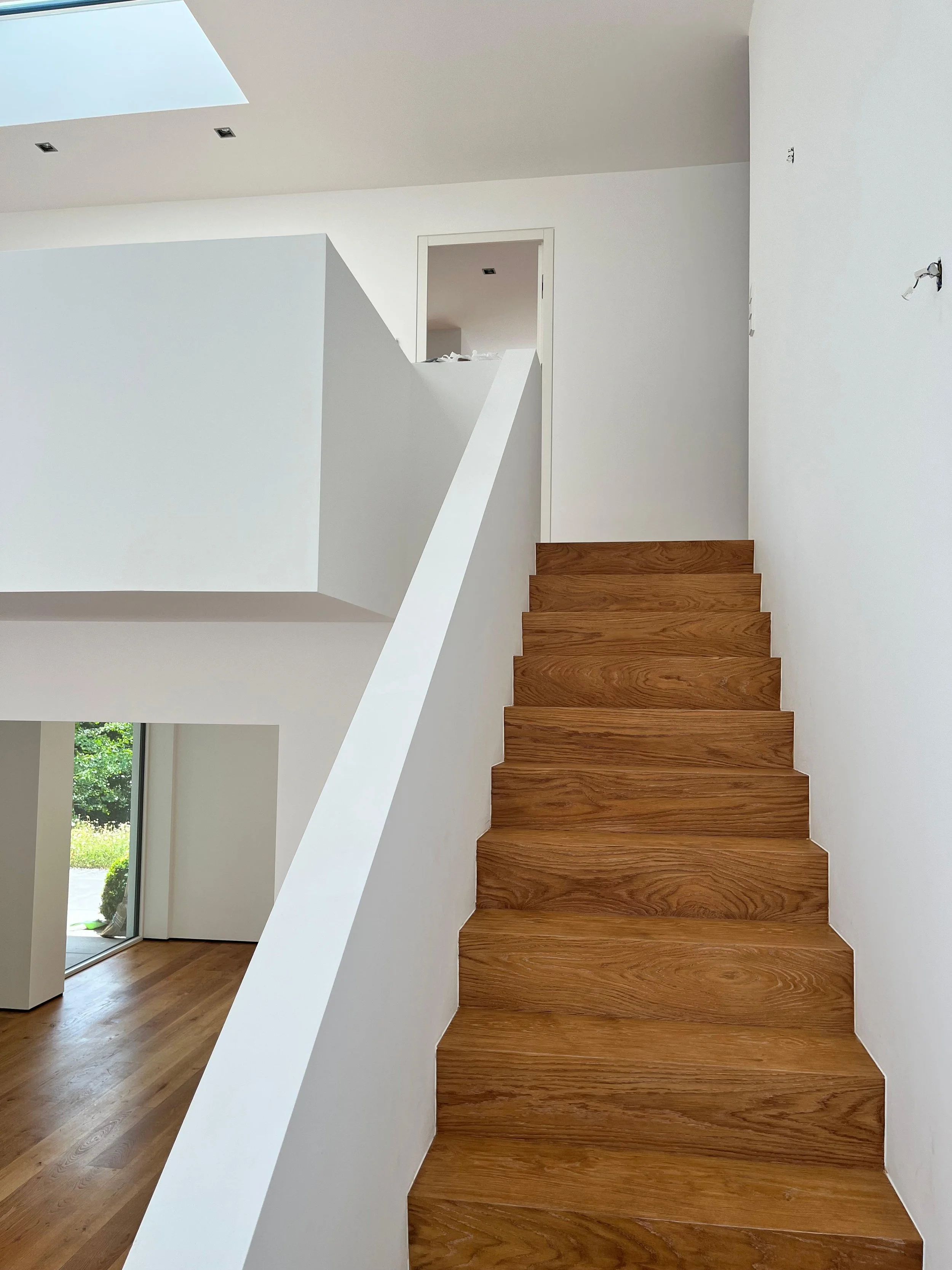 Innenraum eines modernen Hauses mit holzfarbenen Treppen, weißem Wandanstrich und einer großen Dachfensteröffnung, die Tageslicht hereinscheinen lässt.
