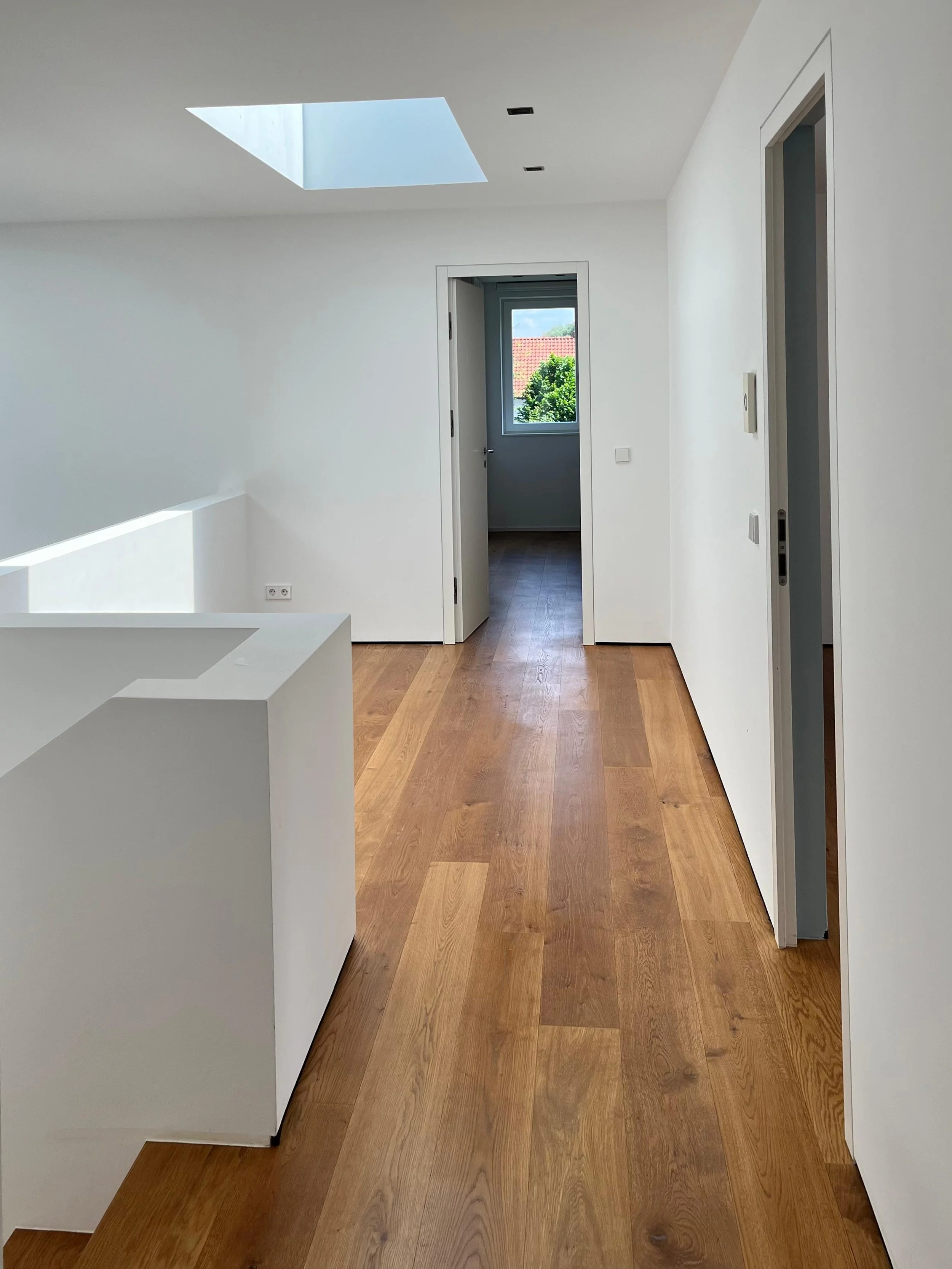 Ein heller, moderner Innenraum mit Holzboden, weißen Wänden und Decke. Es gibt einen offenen Raum mit Türöffnungen, durch die ein Fenster mit Blick auf ein Dach und Bäume zu sehen ist. Helle Tageslichtquelle von oben.