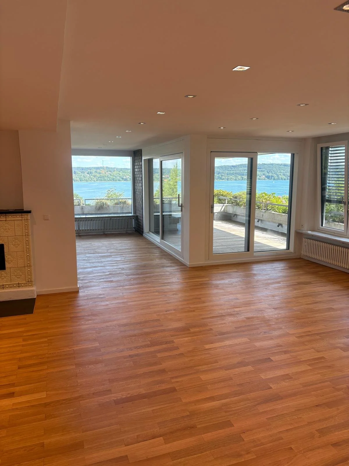 Leeres Wohnzimmer mit Holzfußboden, große Fenster und Balkon mit Blick auf einen Wasserarm, grüne Hügel und Bäume in der Ferne.
