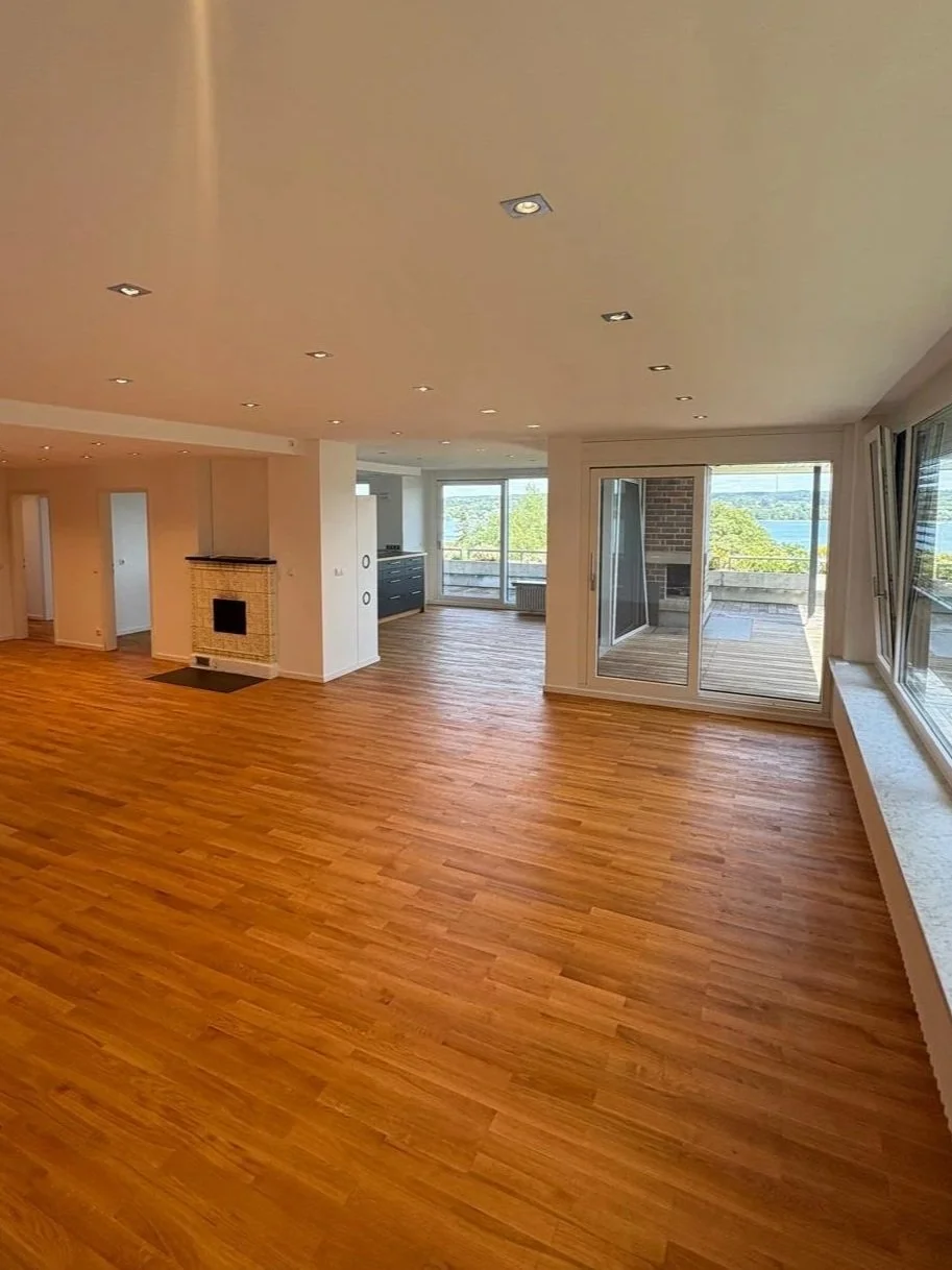 Ein helles, modernes Wohnzimmer mit Holzboden, großen Fenstern und einem Balkon, Blick nach draußen auf die Natur.