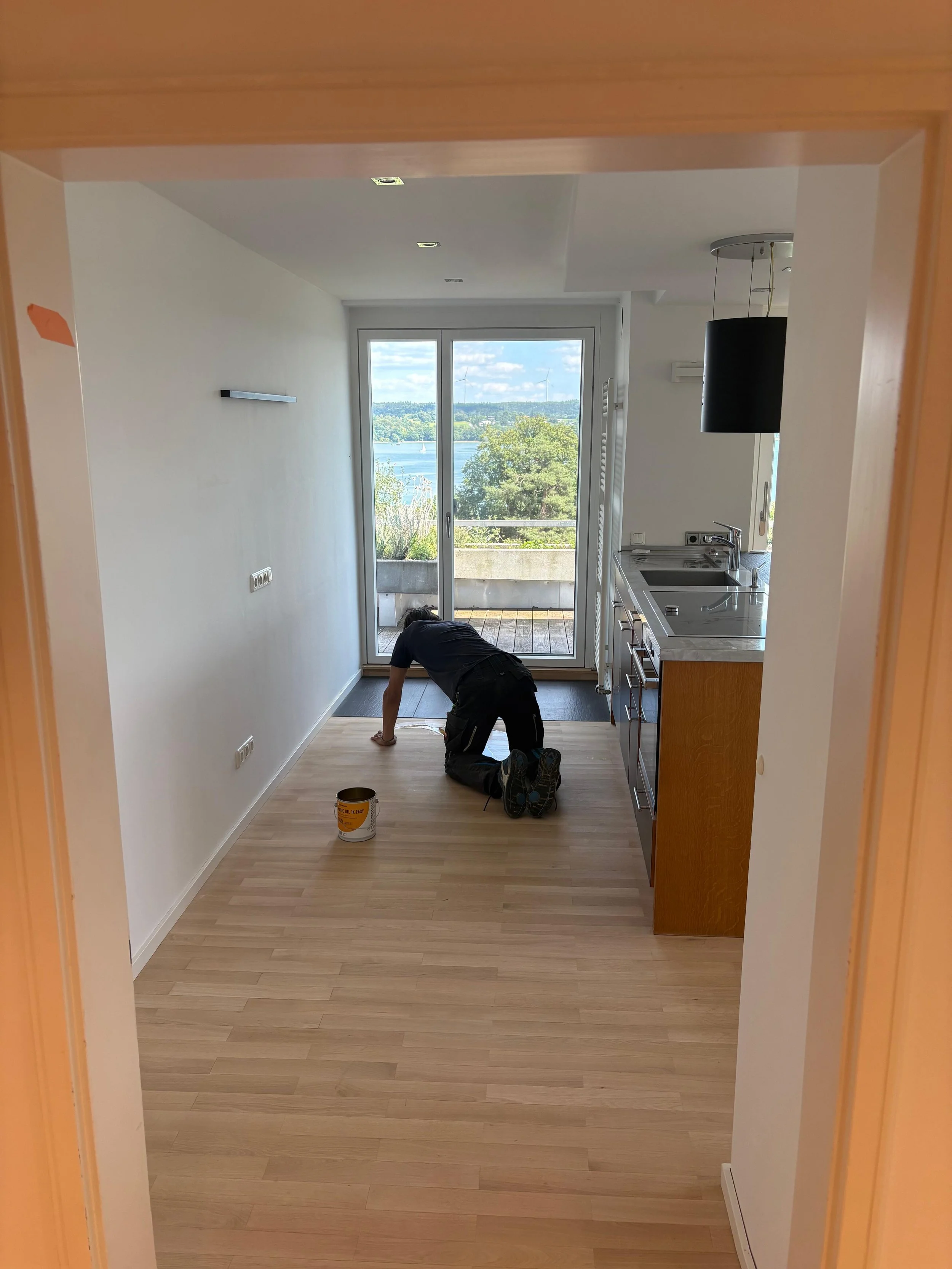 Ein Mann arbeitet auf Knien auf dem Holzfußboden in einer modernen Küche, während er an der Wand streicht oder verfugt. Im Hintergrund sieht man große Fenster mit Blick auf einen Fluss und grünen Büsch. Es ist Tag, das Wetter ist sonnig und klar.