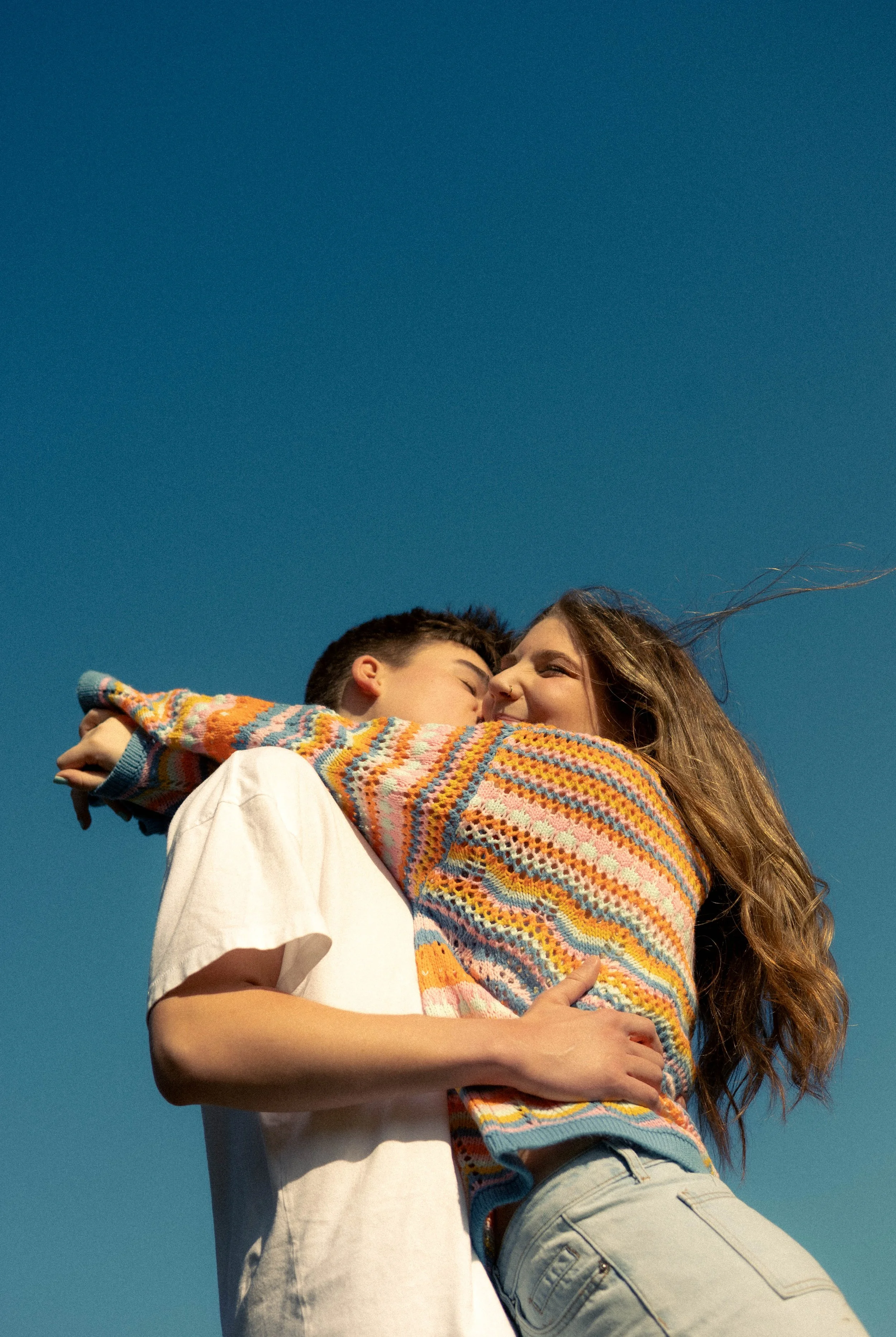 A young couple embracing and kissing outdoors against a clear blue sky.