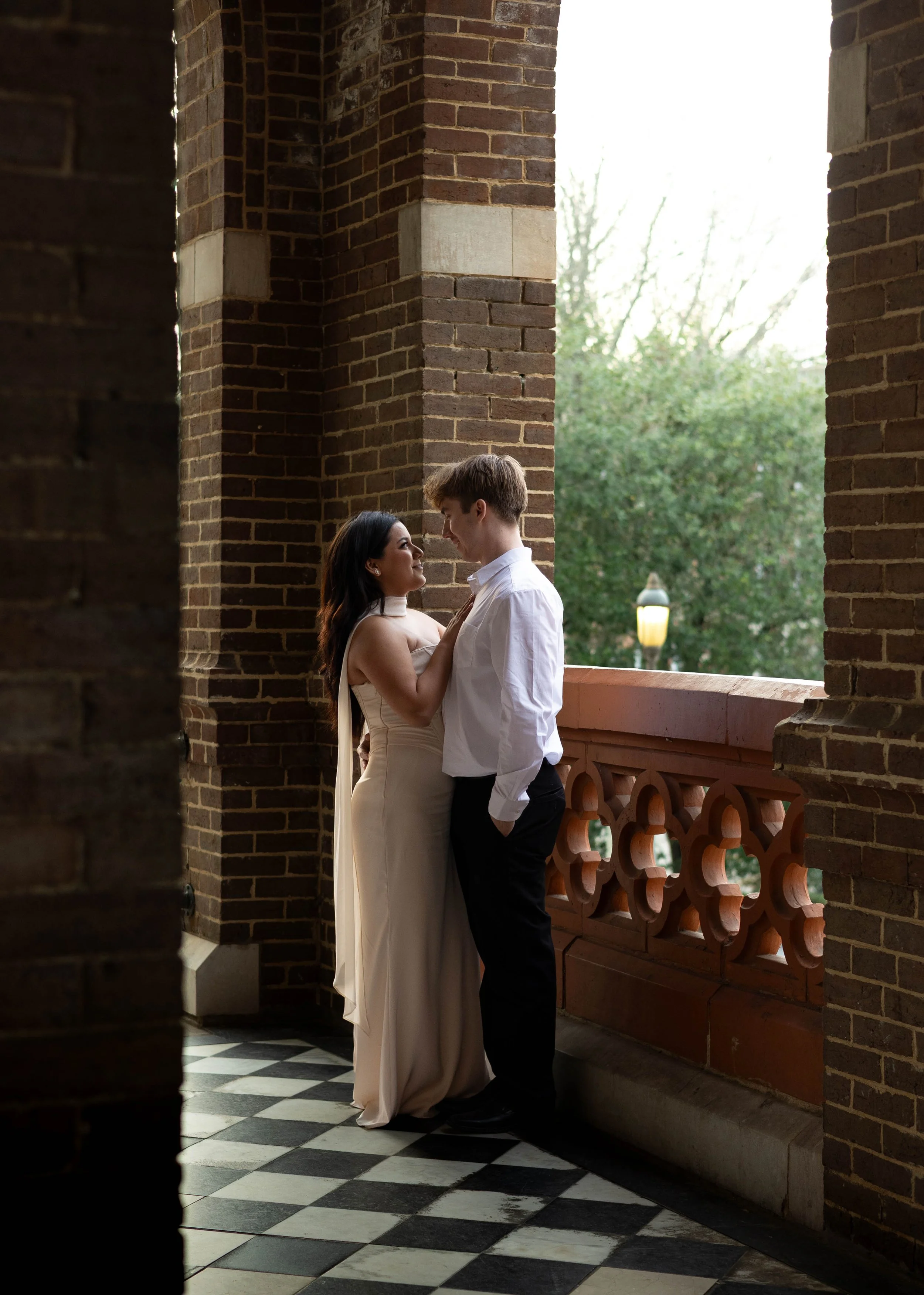 A couple stands close together on a balcony with brick walls and a checkerboard floor. The woman wears a light-colored, strapless dress, and the man wears a white shirt and black pants. They gaze into each other's eyes, with the woman's hand on the man's chest. The background shows greenery and a streetlamp outside.