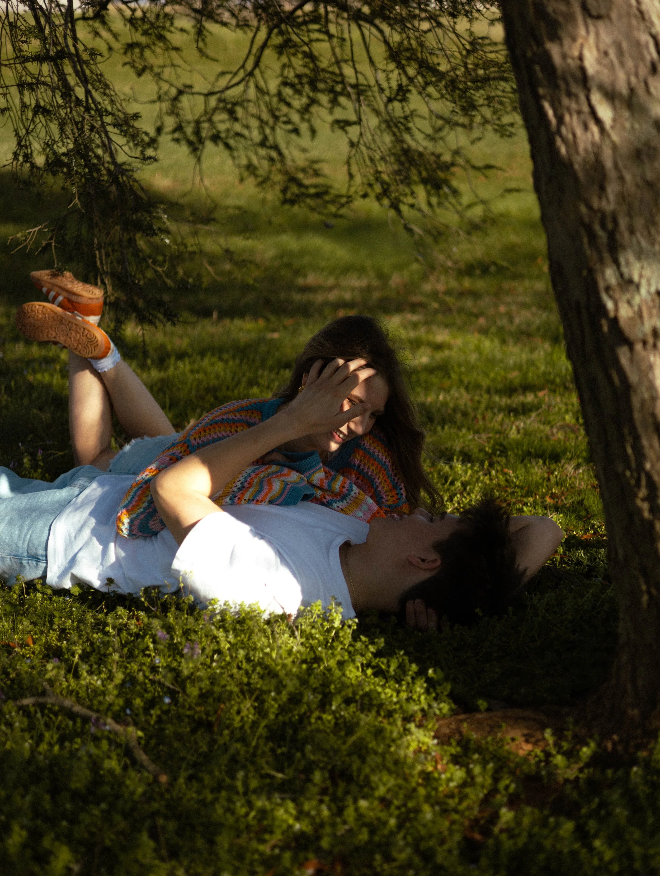 A young couple lying on the grass under a tree, looking at each other, with the girl touching the boy's face.