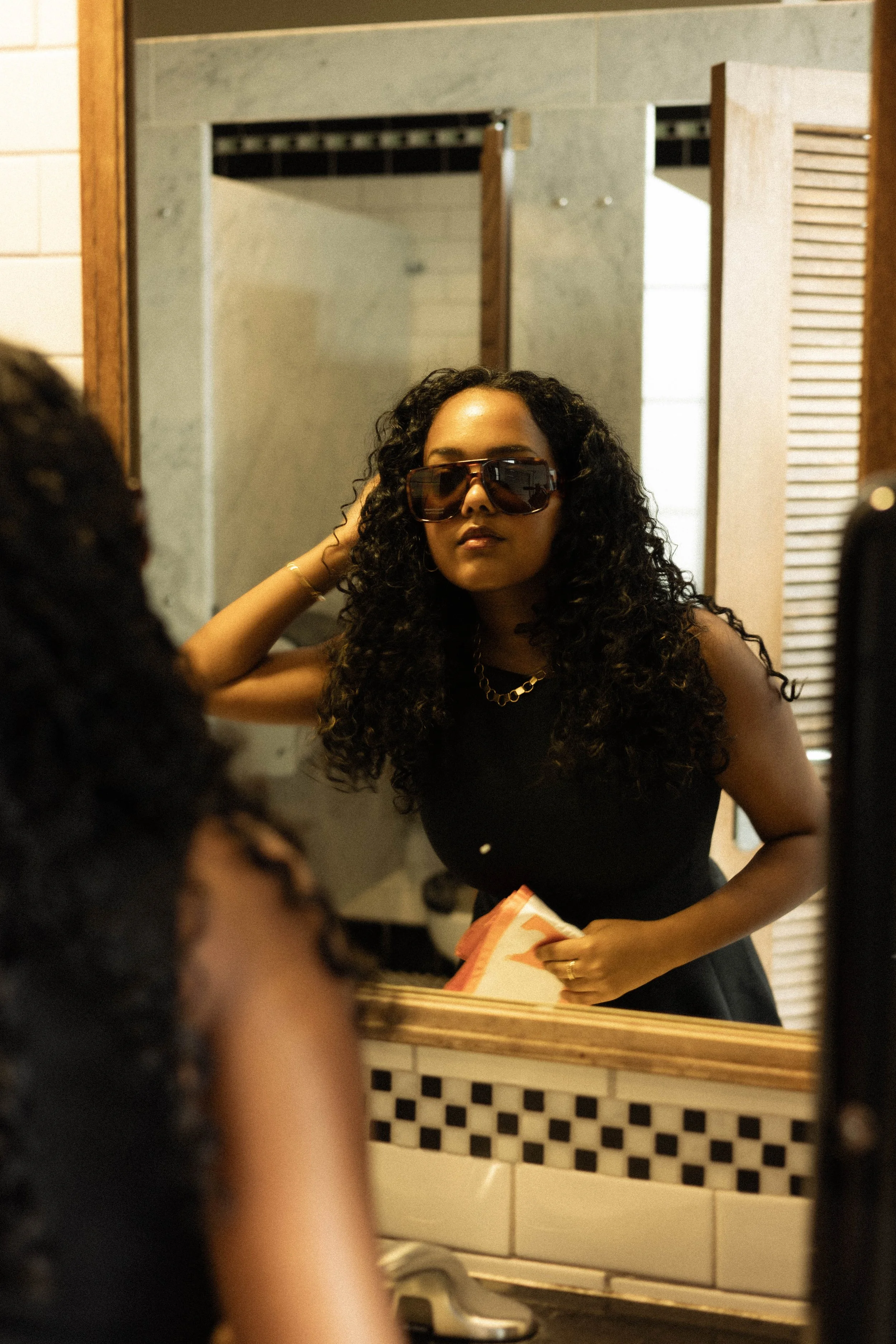 A woman with curly hair wearing sunglasses, a black outfit, and a gold necklace is looking in the mirror in a bathroom.