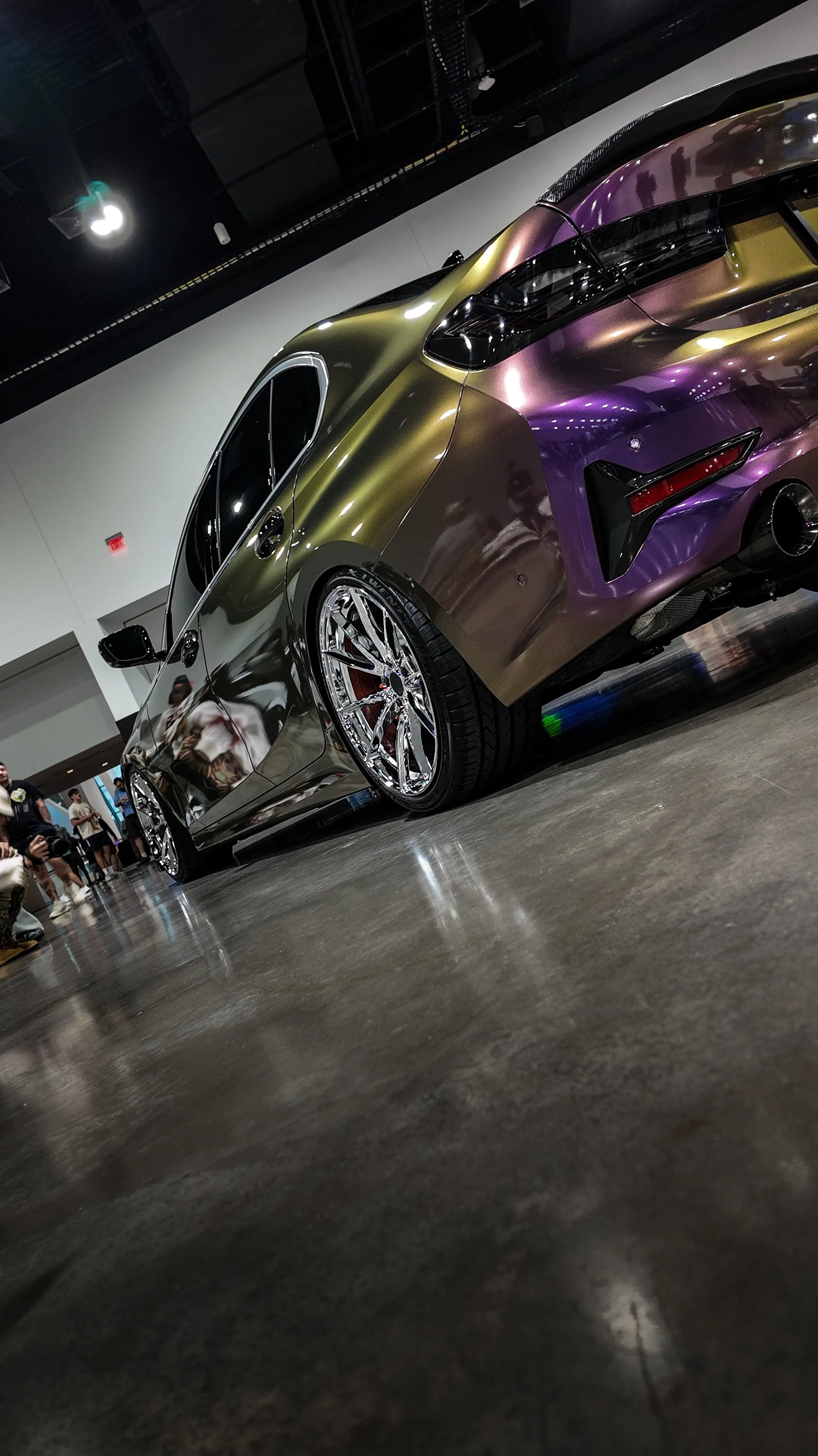 Low-angle view of a customized sports car with a chameleon-style paint job, chrome wheels, and large tires at an indoor car exhibition.