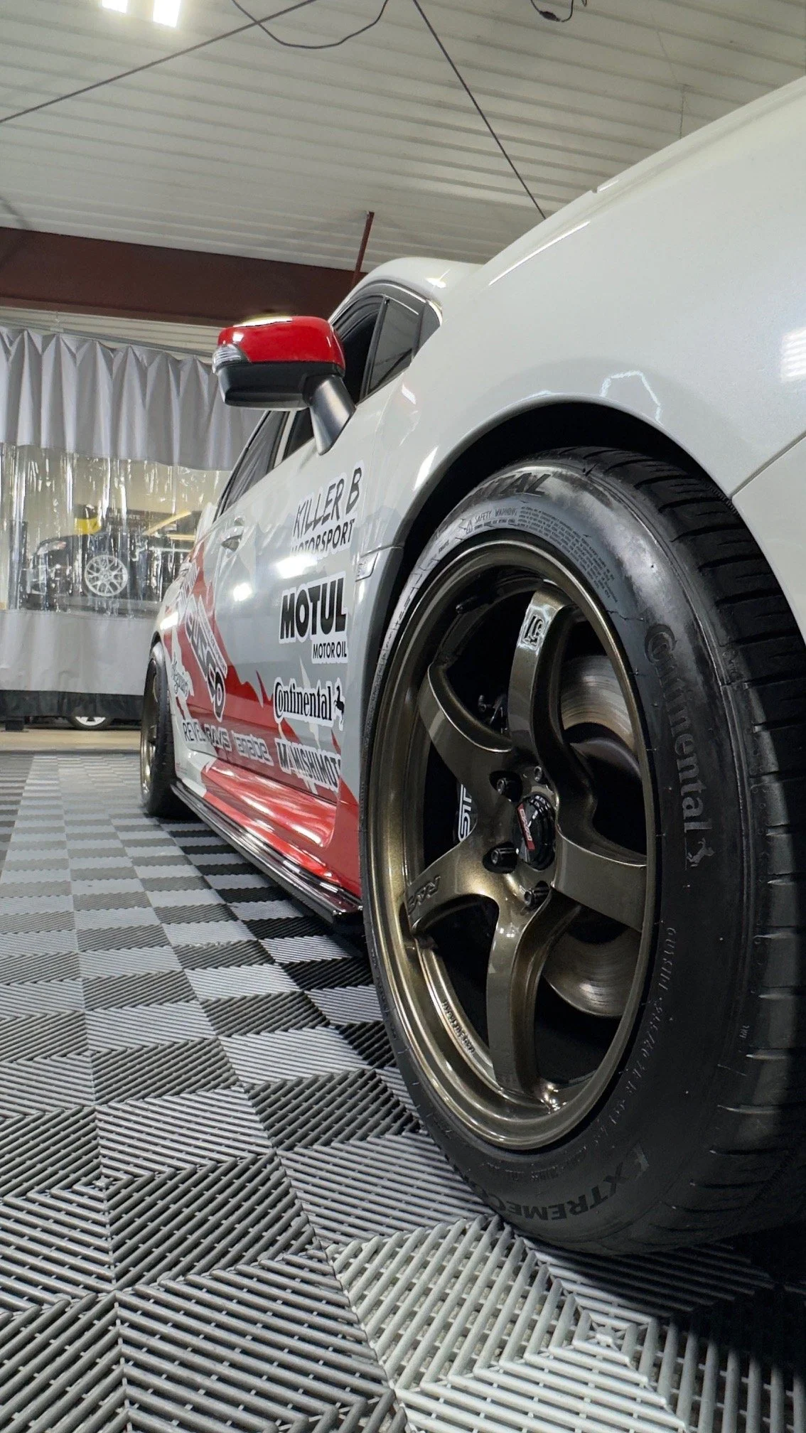 A race car with sponsor decals, close-up of the front tire and wheel on a checkered floor, indoor setting.