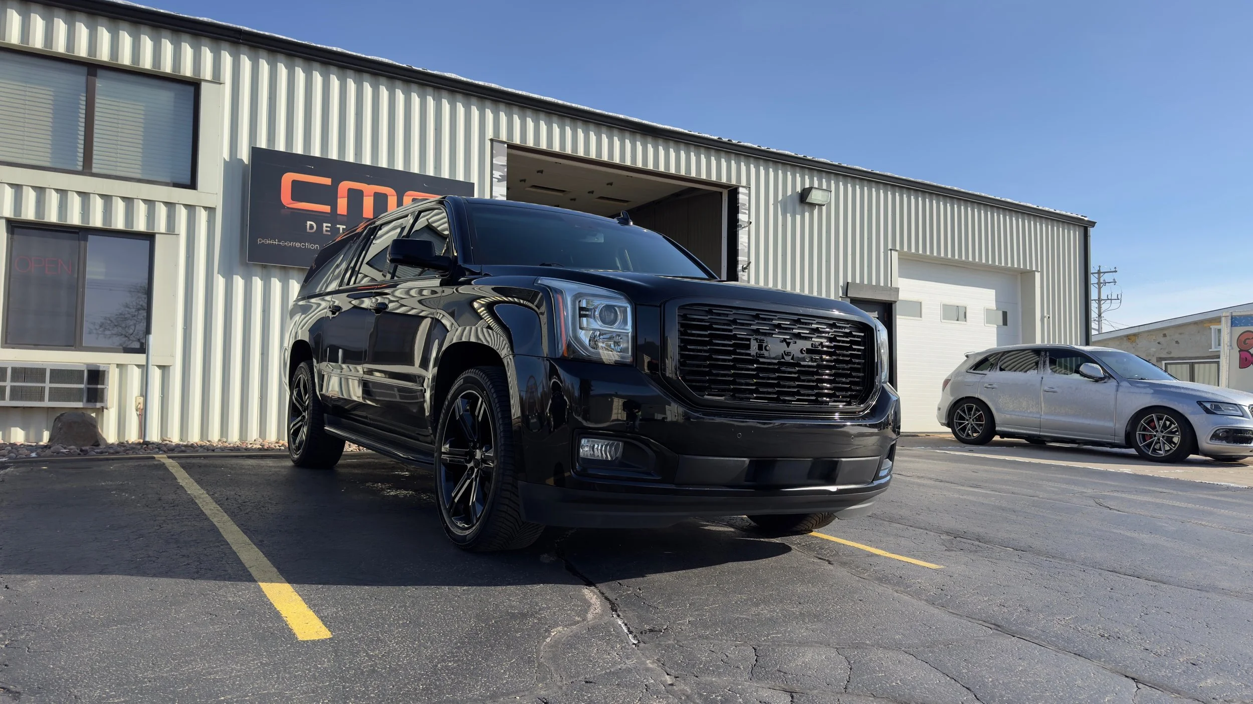 Black SUV parked in front of a building with a sign that reads 'CMC Detailing'. A silver car is parked nearby in a parking lot.