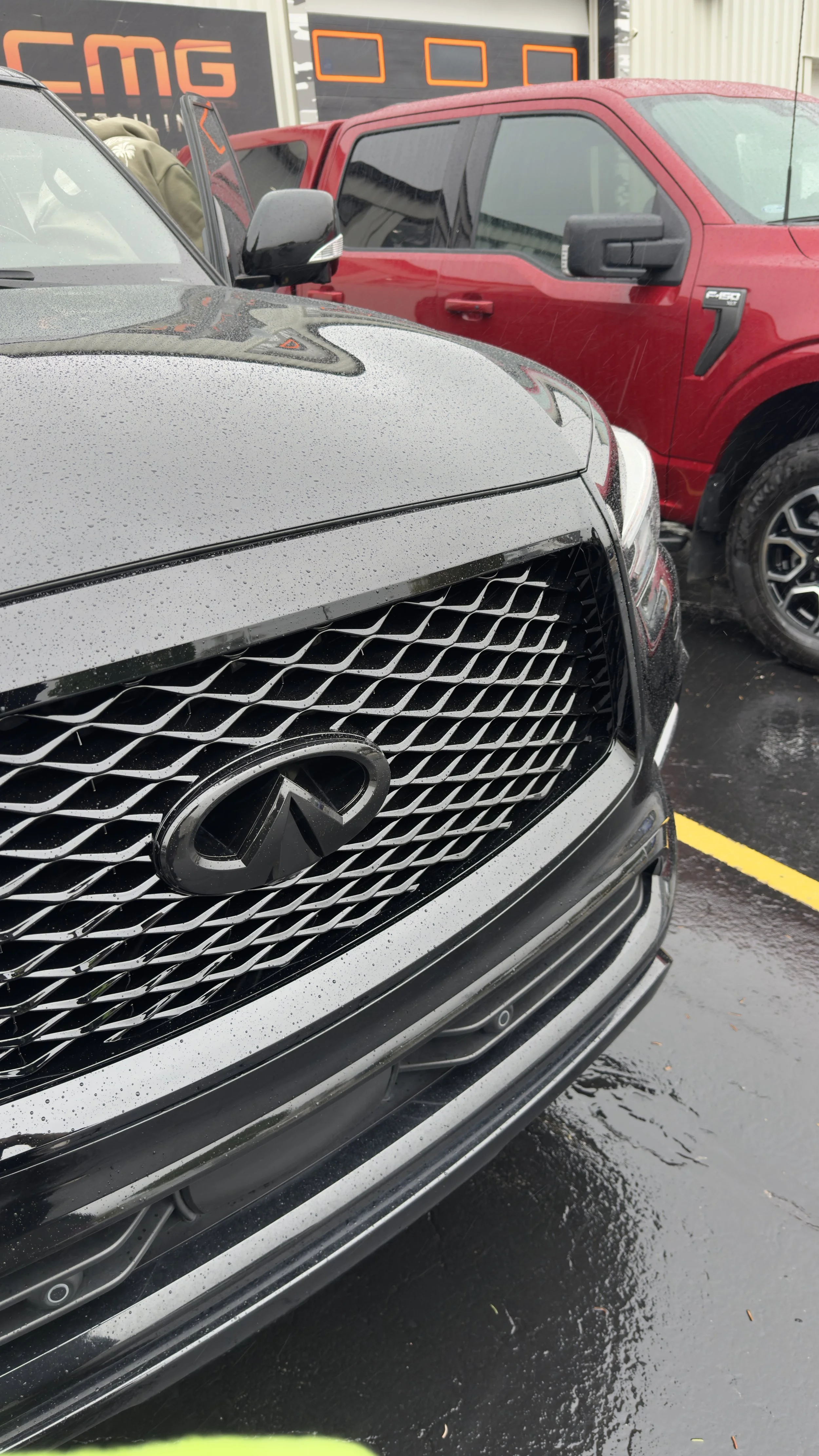 Front view of a black Infiniti vehicle with raindrops on its surface, parked in front of a red truck and a building with a sign that reads 'EMG' on a rainy day.