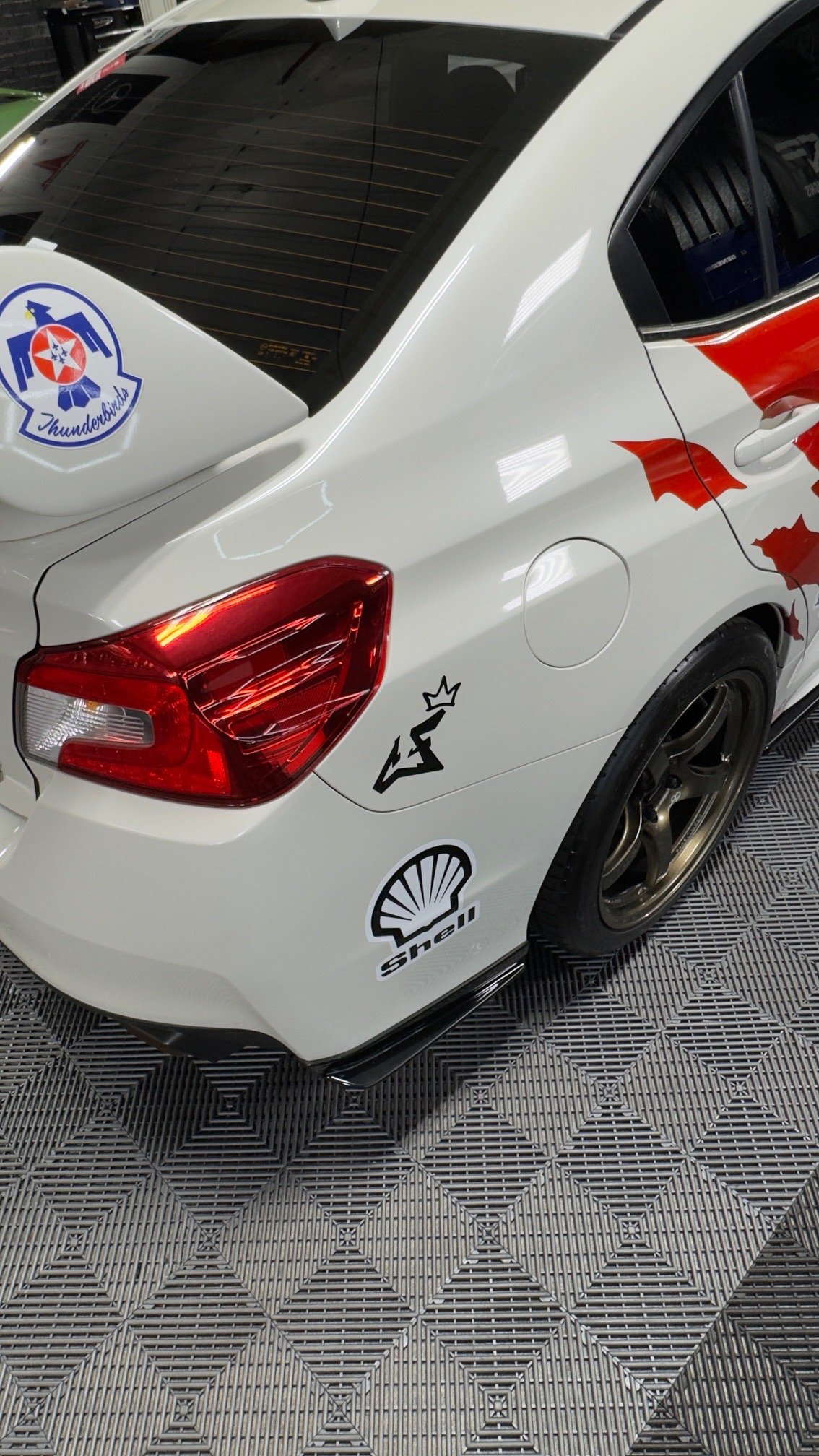 Close-up of a white race car with various stickers, including Shell, a crown illustration, and a logo with a bird, parked on a patterned floor.