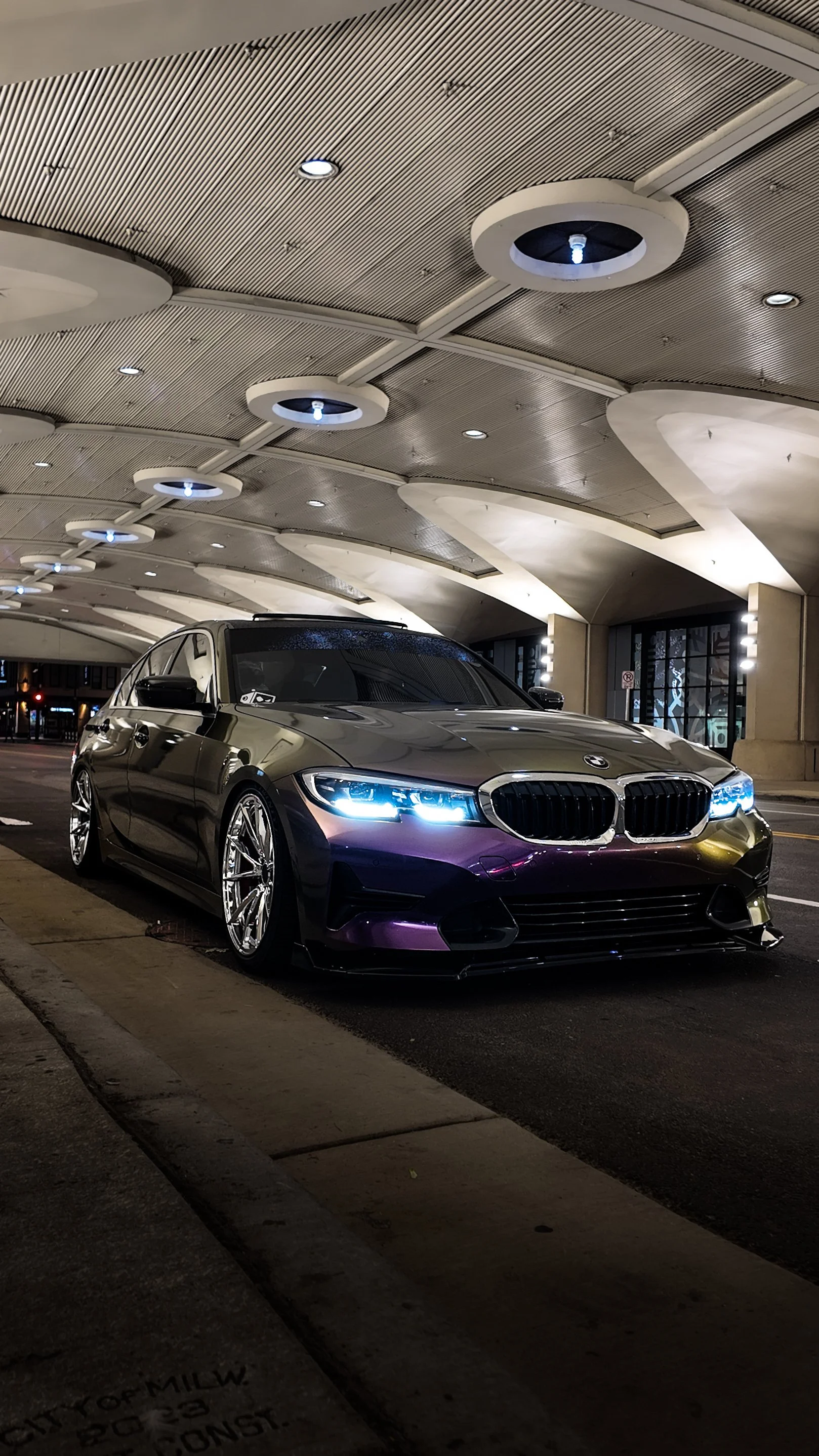A black BMW sedan parked under an architectural canopy at night, with illuminated blue headlights and reflective surface.