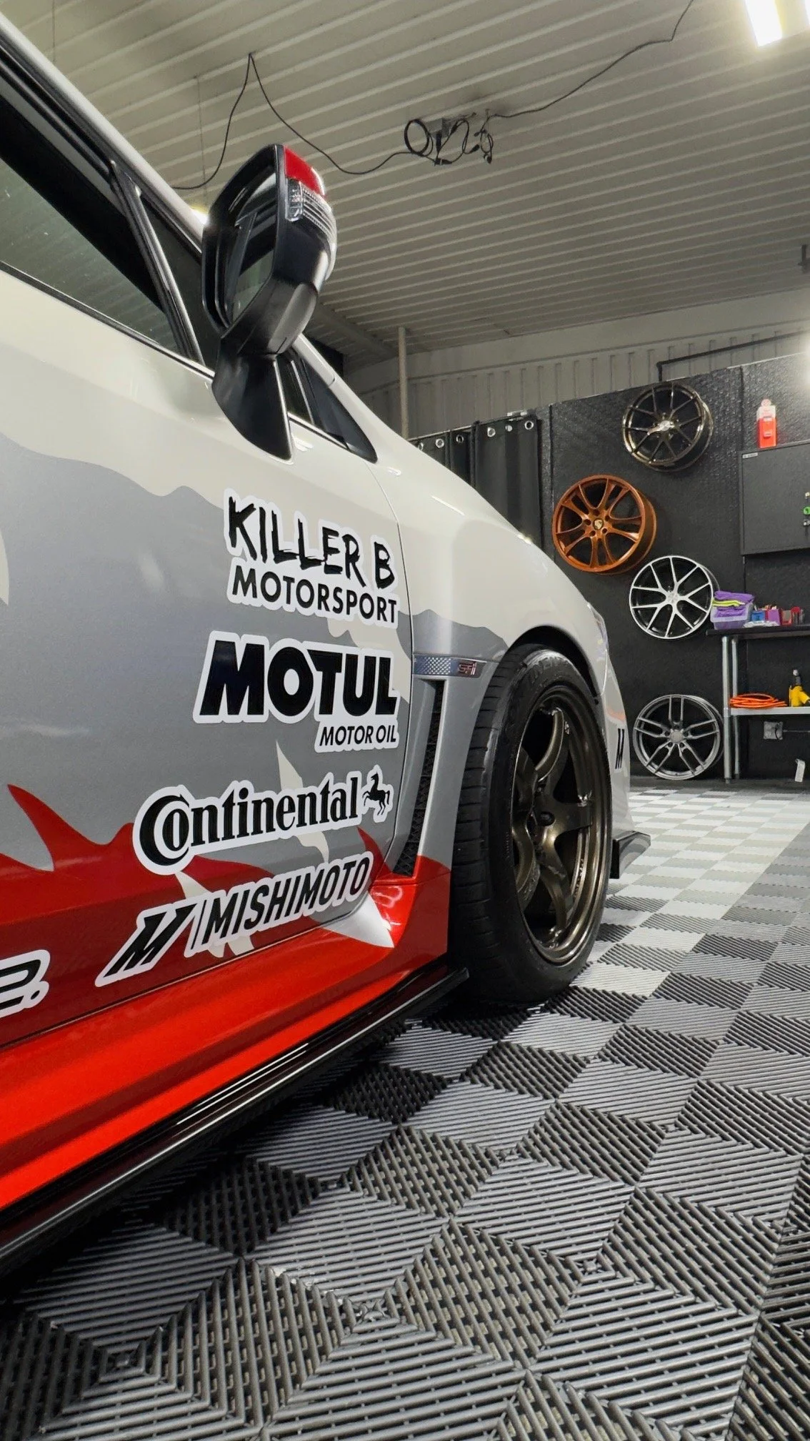 Close-up of a race car with sponsor decals, inside a garage with multicolored alloy wheels mounted on the wall and tools on a workbench.