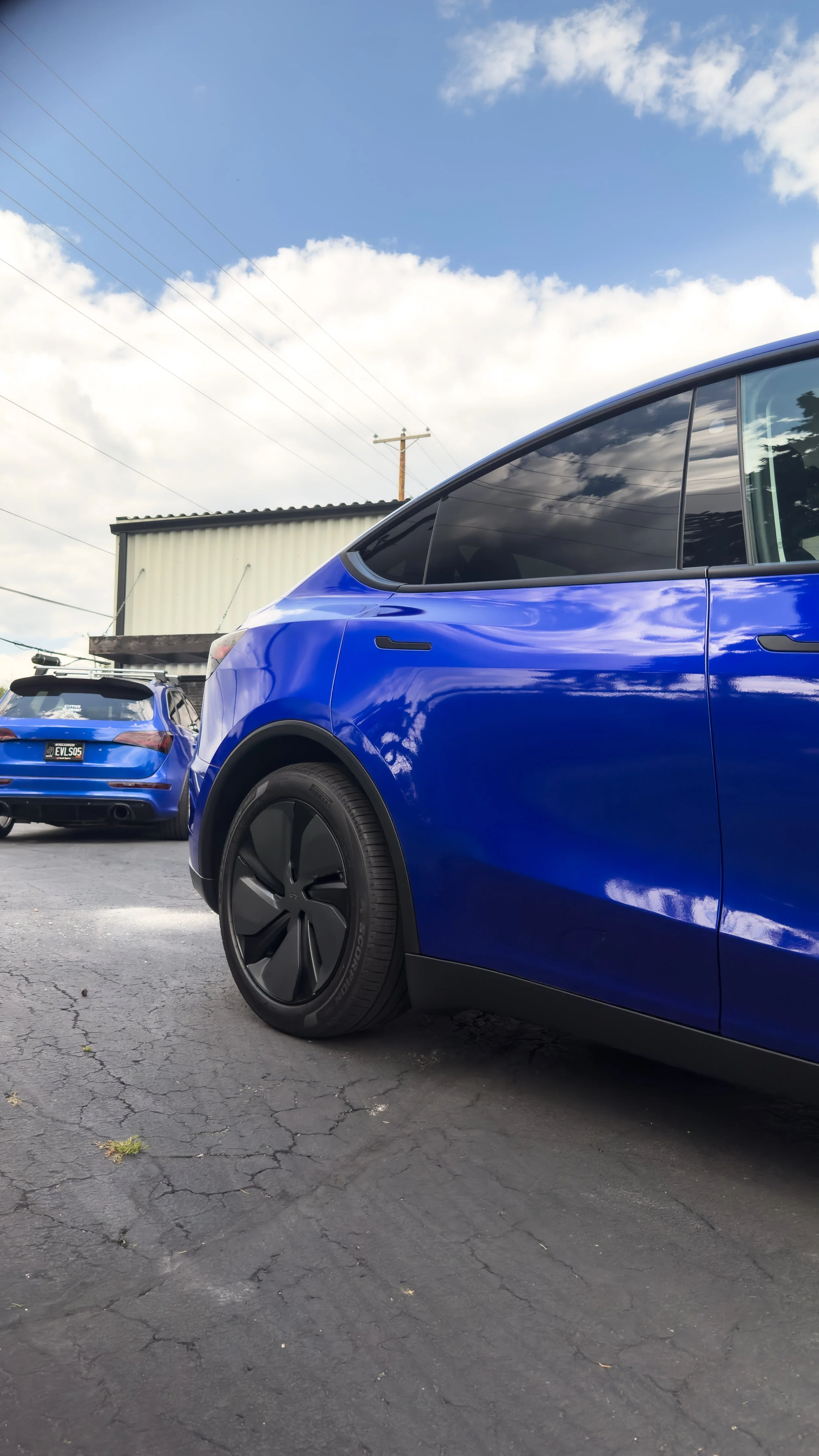 Close-up of a blue electric vehicle parked on cracked asphalt with another blue car in the background, a shed, power lines, and a partly cloudy sky.