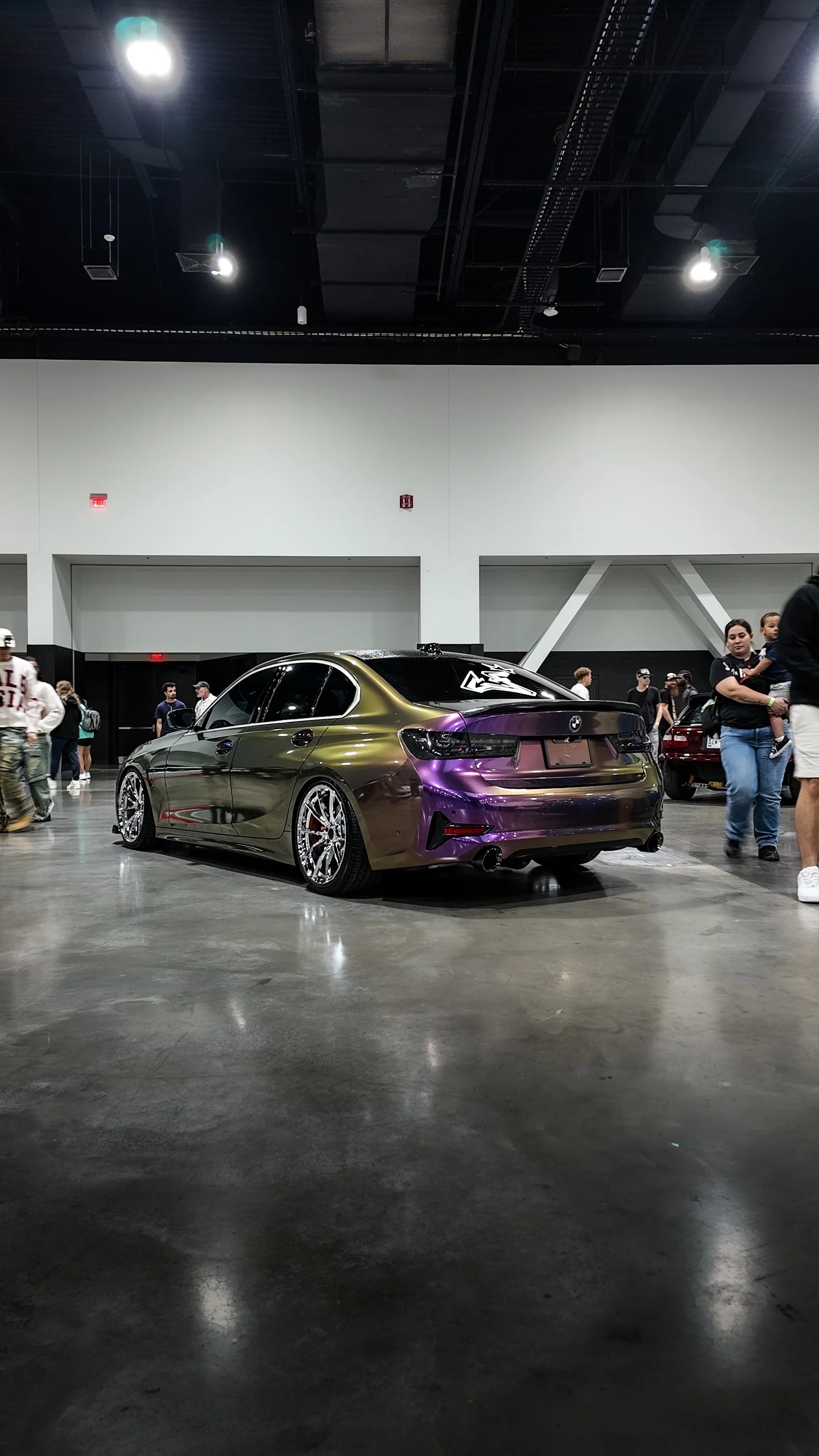 A customized BMW sedan with unique multicolor reflective paint at an indoor car show, with people walking around and viewing other vehicles.