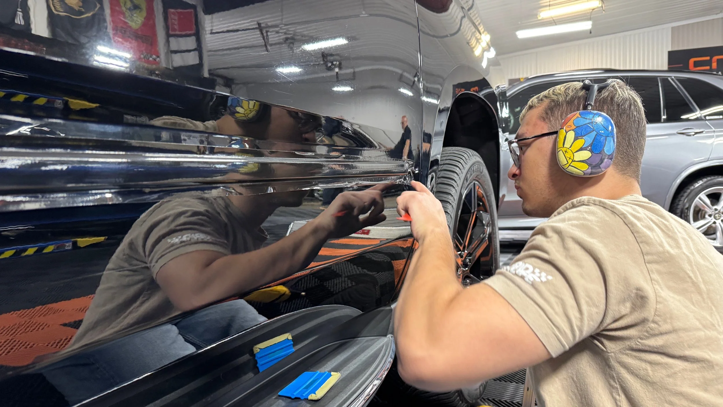 A young man wearing a beige shirt, glasses, and colorful floral earmuffs is working on a black car in a garage. He is using a tool on the car's surface, and his reflection can be seen on the car's shiny exterior. There are two other cars visible in t