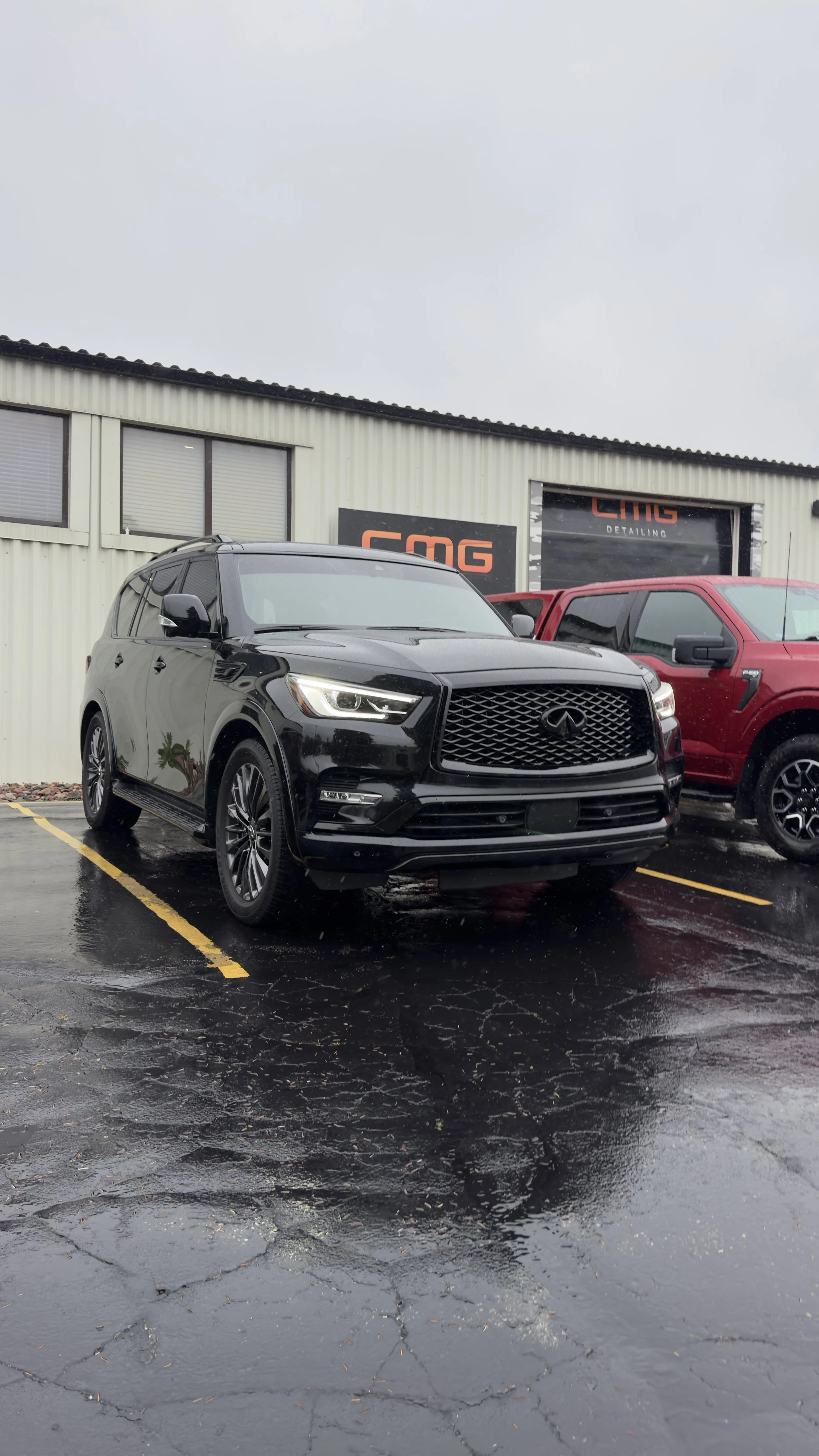 Black SUV parked on wet pavement in front of a building with a sign reading 'CMG Detailing' and red vehicle beside it, on a rainy day.