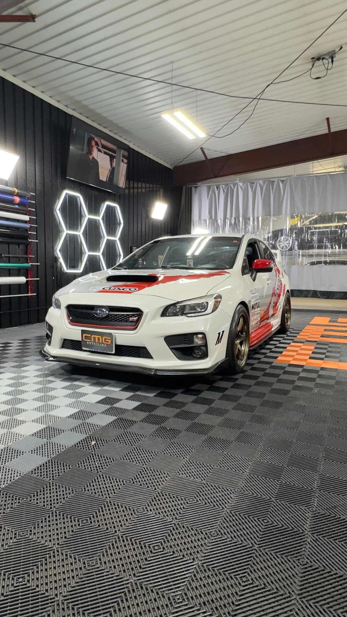 White race car with racing decals inside an indoor garage with black and orange checkered flooring, illuminated hexagon wall lights, and a wall-mounted TV.