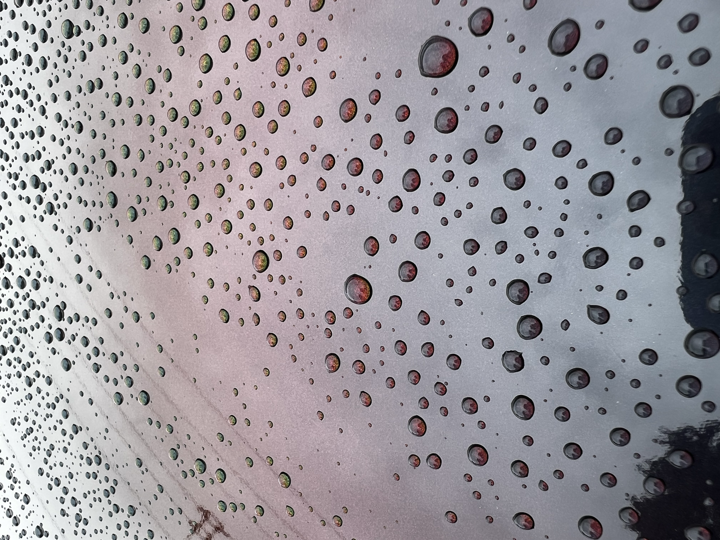 Raindrops on a car window reflecting a cloudy sky with a hint of red and green trees.