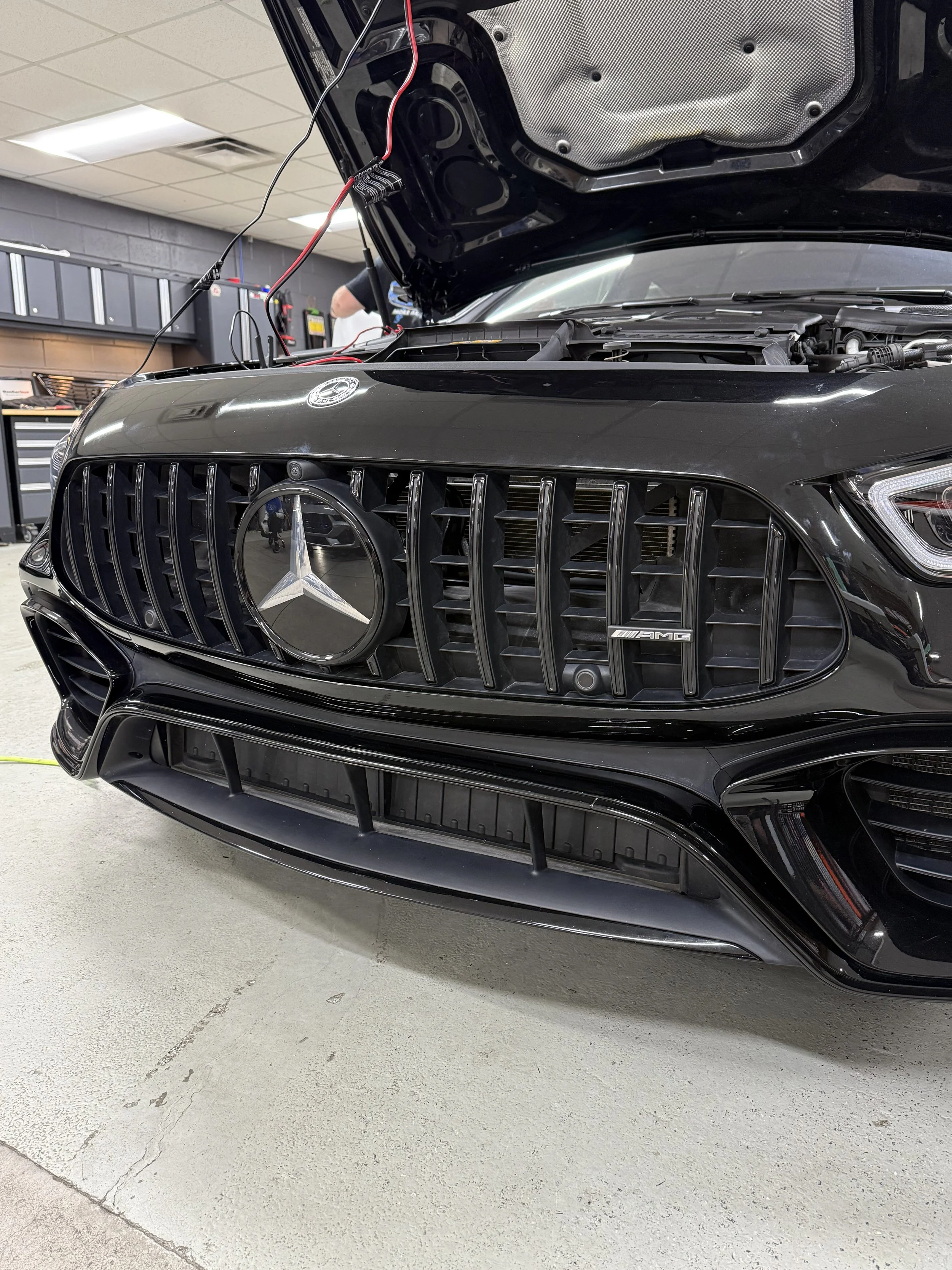 Front view of a black Mercedes-Benz car with its hood open, showing the grille and emblem, in a garage or workshop.