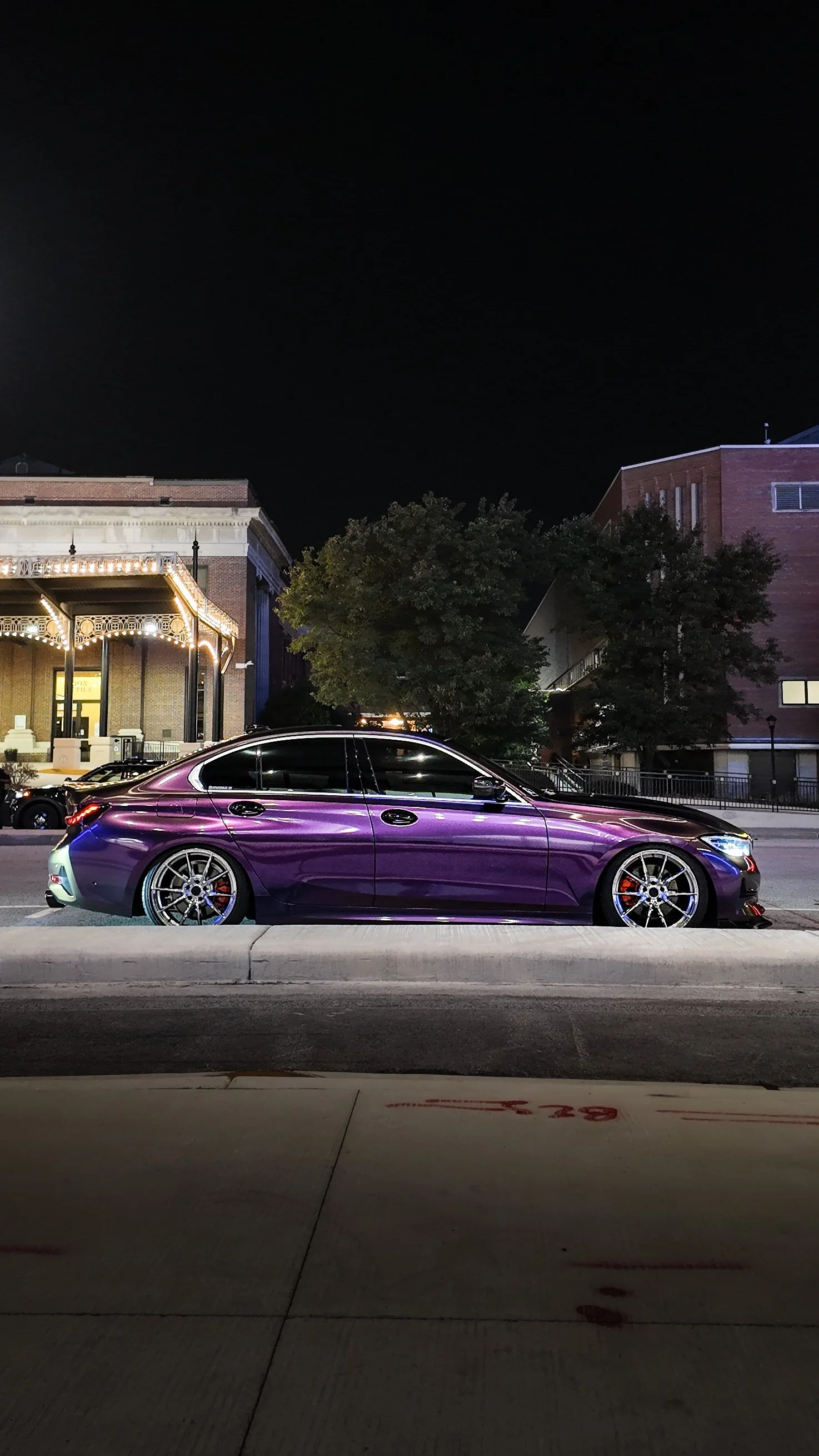 A purple luxury sedan parked on a city street at night, with buildings and trees in the background.