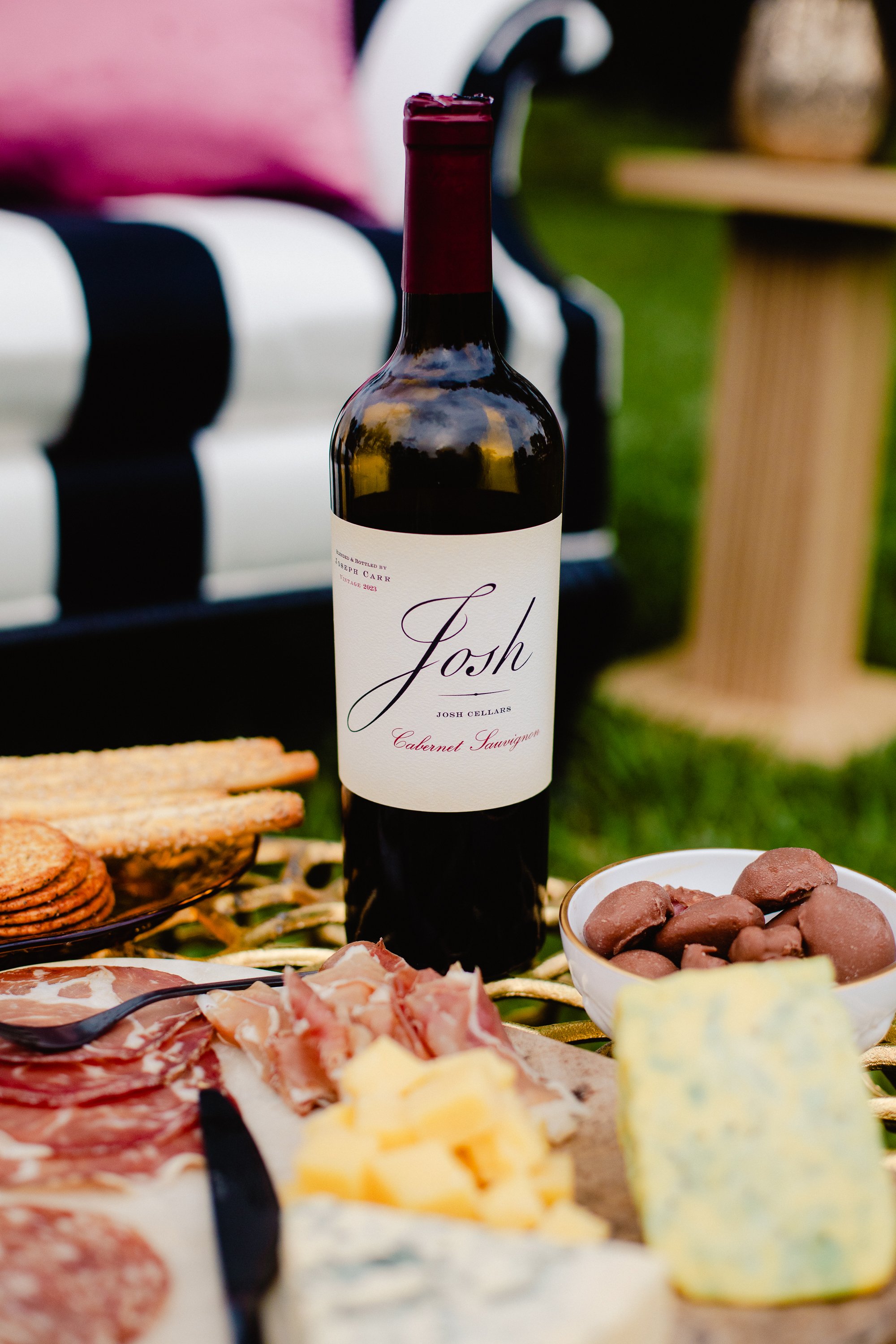 A bottle of Josh Cellars Cabernet Sauvignon wine on a table with a charcuterie board, cheese, and chocolates, set outdoors.