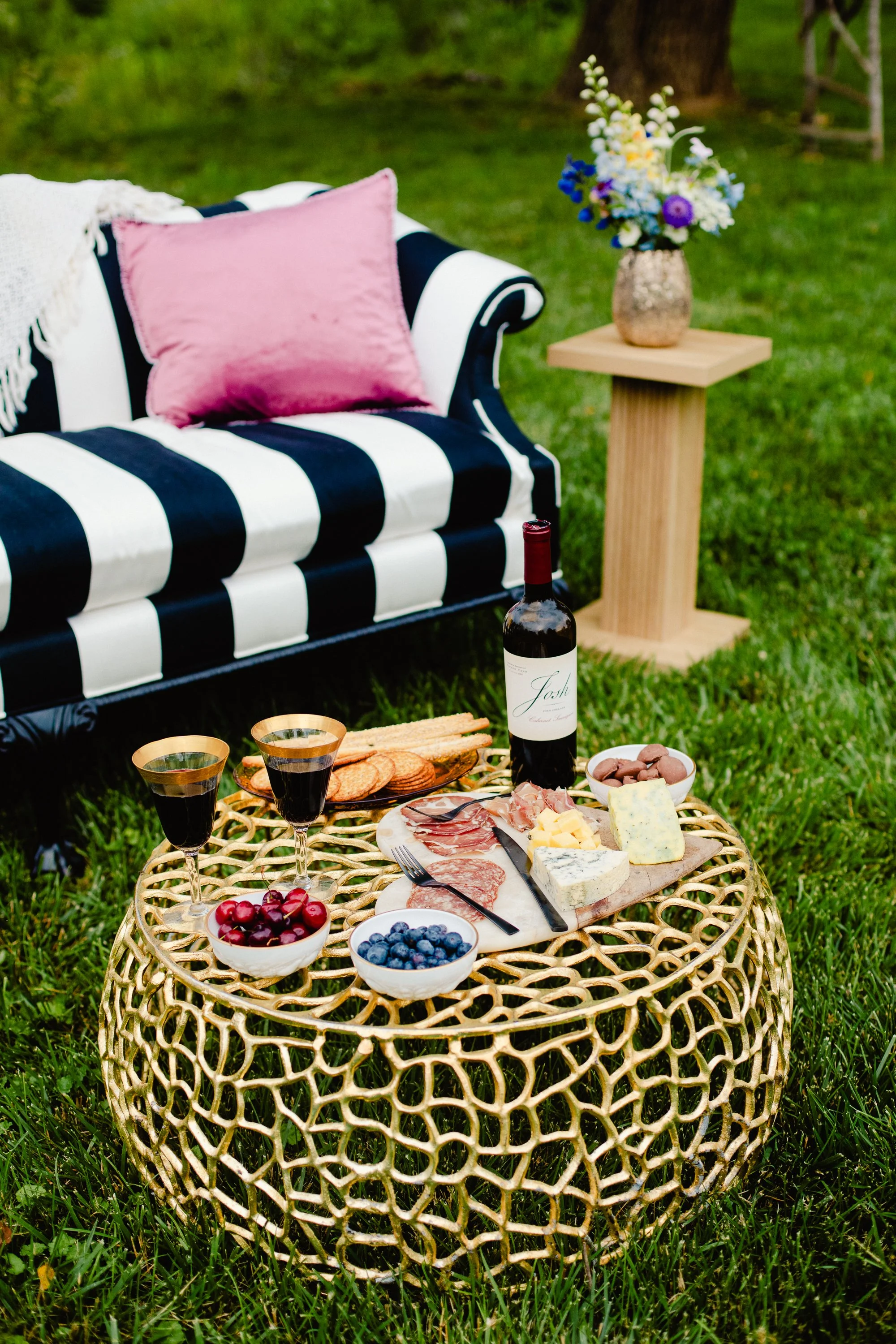 Outdoor picnic setup with a round woven table holding cheese, meats, grapes, blueberries, and cherries, along with wine glasses filled with red wine. In the background, there is a black and white striped sofa with a pink pillow and a side table with 