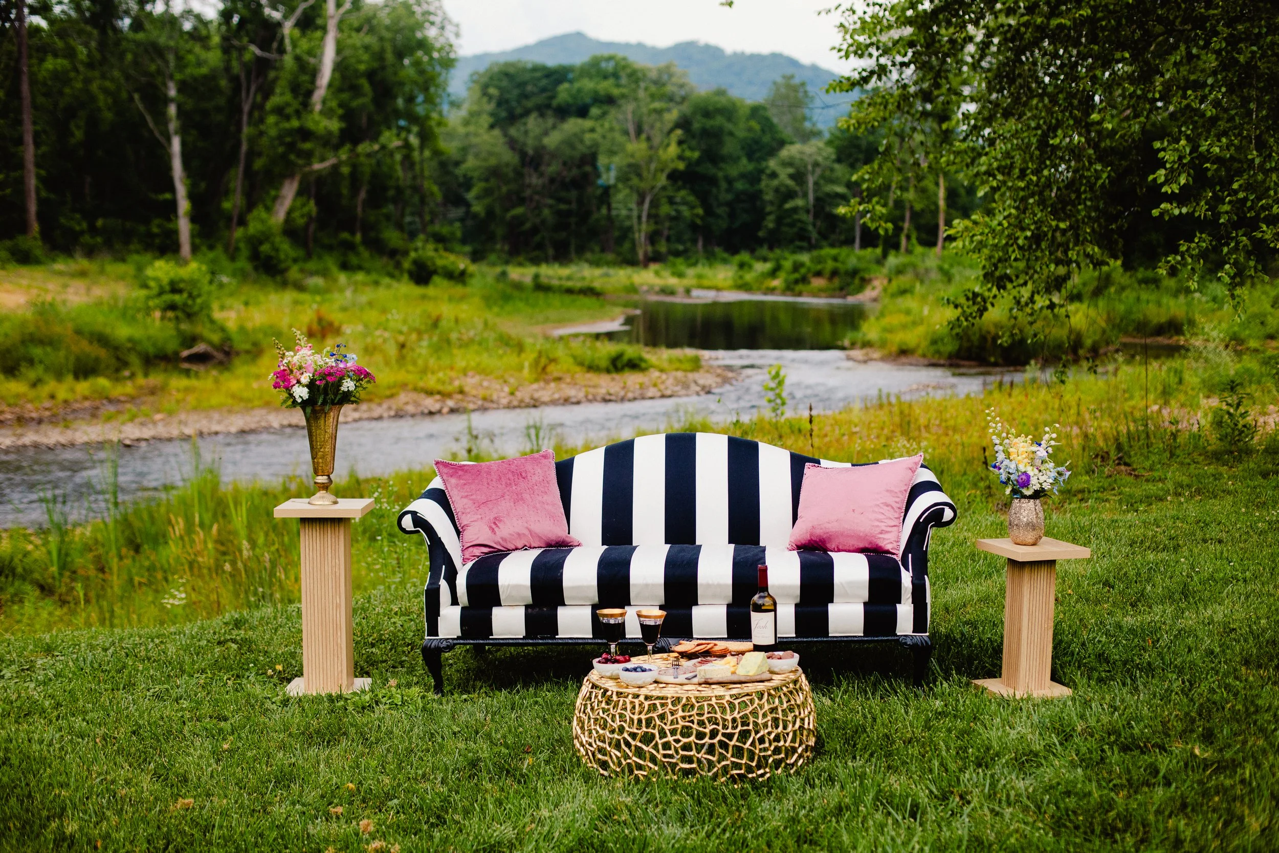 A black and white striped sofa with pink pillows, set outdoors in a lush green landscape near a river, with side tables holding vases of colorful flowers, a bottle of wine, glasses, and a cheese platter on a woven table in front.