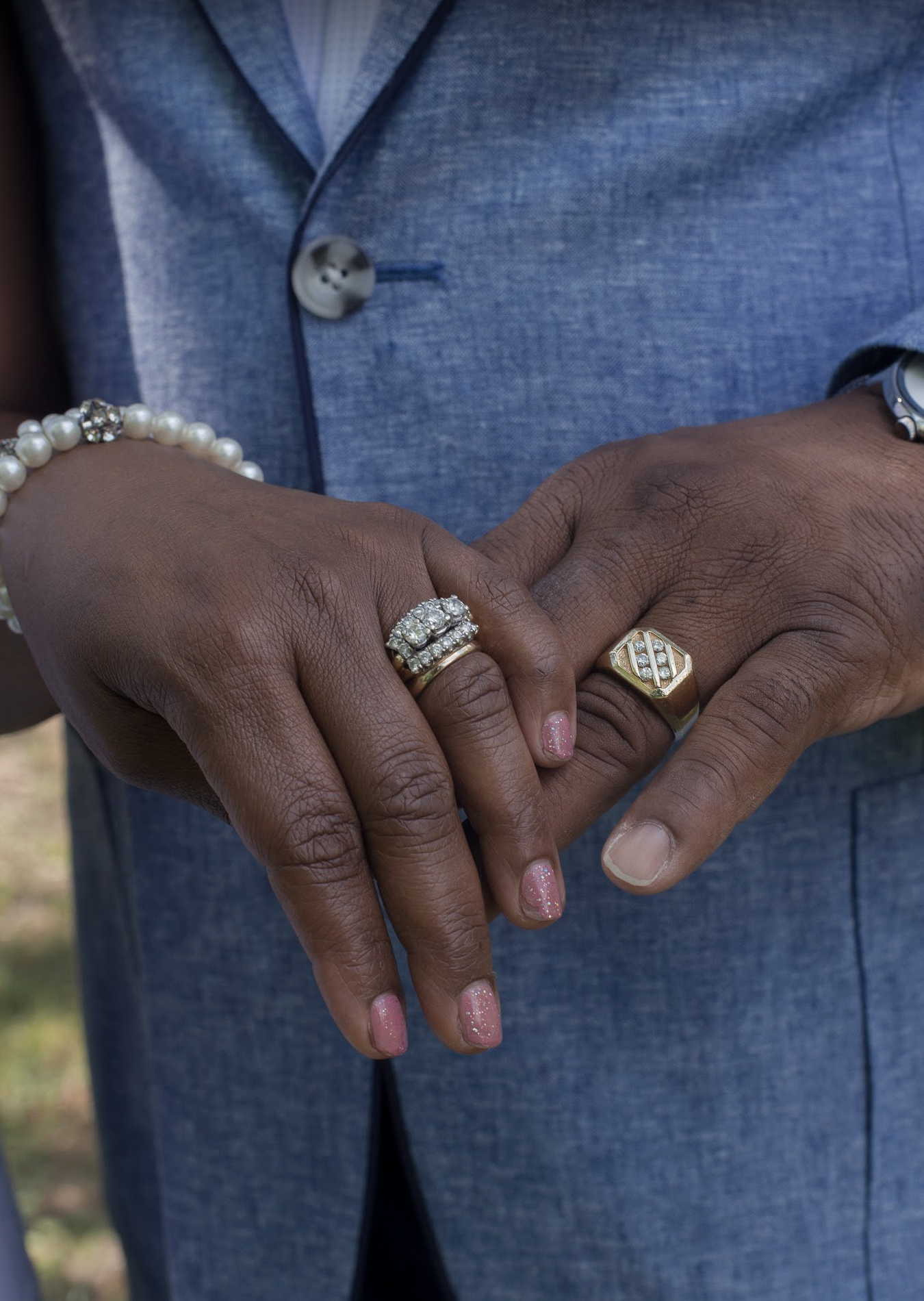 Close-up of two hands with rings and jewelry, one hand wearing a pearl bracelet and a diamond ring, the other with a gold ring with diamonds, held against a man's chest in a blue suit.