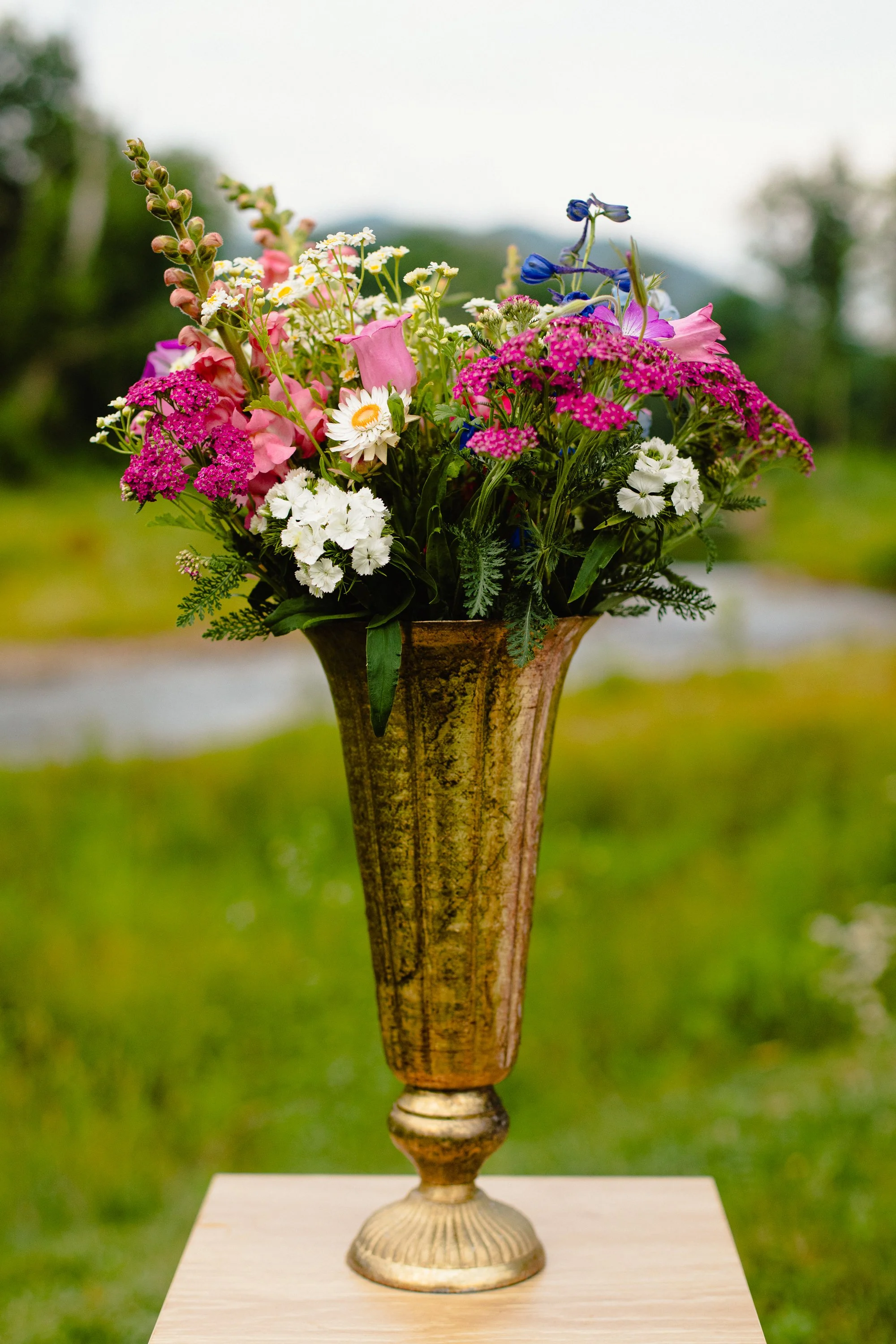 A vase with a floral arrangement of pink, white, purple, and blue flowers placed on a table outdoors with blurred greenery in the background.