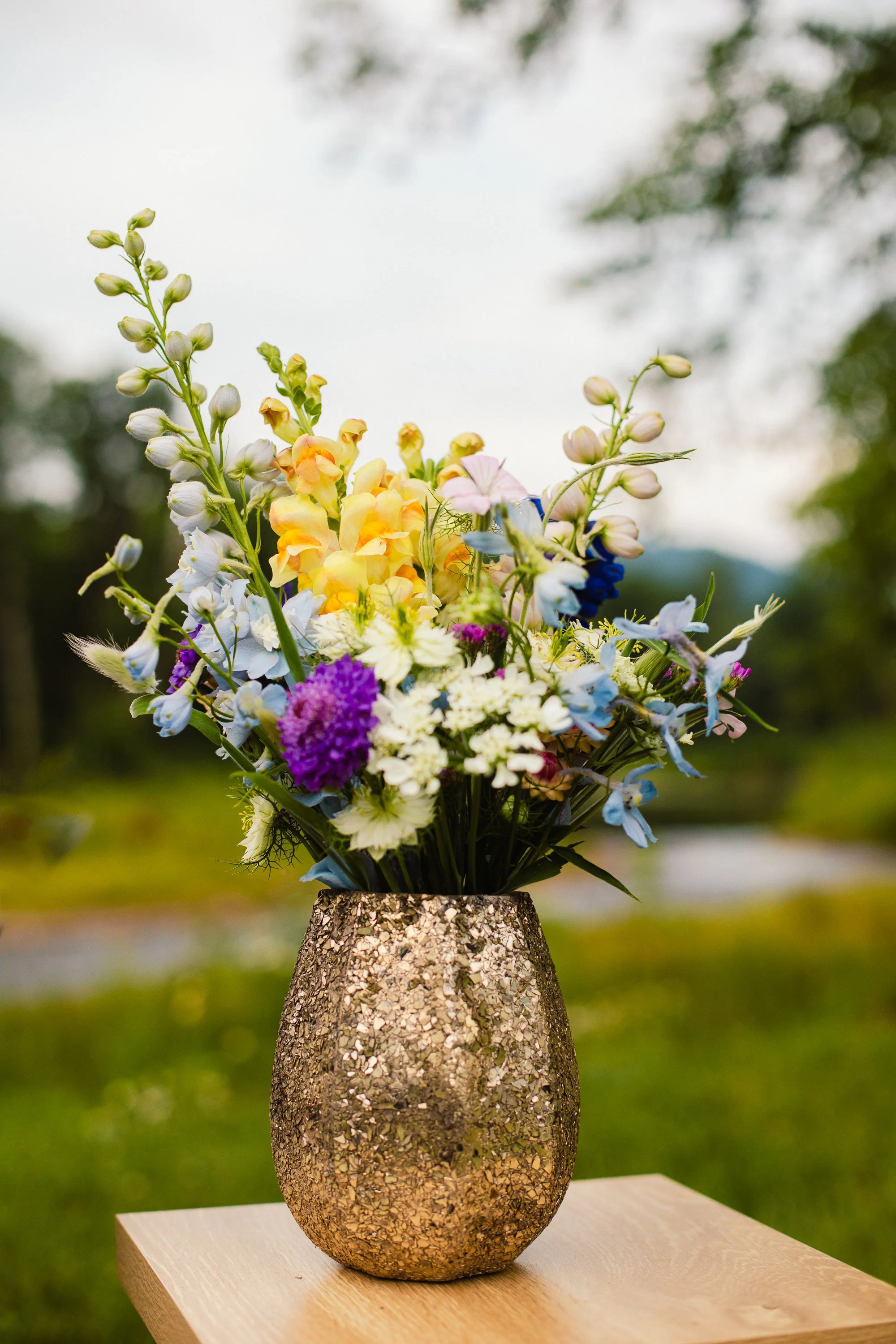 A colorful flower arrangement in a textured stone vase on a wooden table outdoors, with a blurred background of trees and sky.