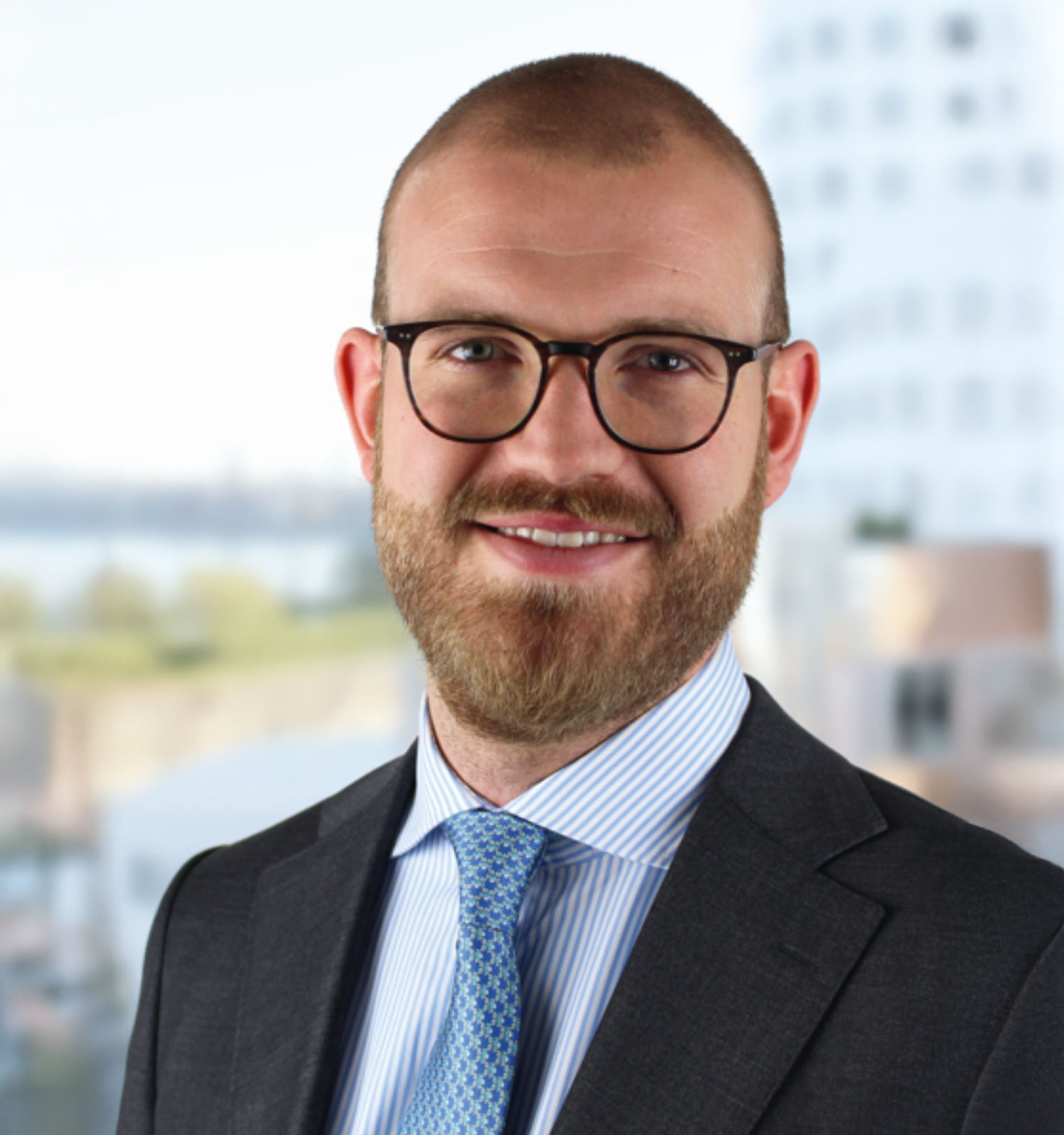 Professional headshot of a smiling man with glasses, a beard, wearing a suit and tie, with a cityscape background.