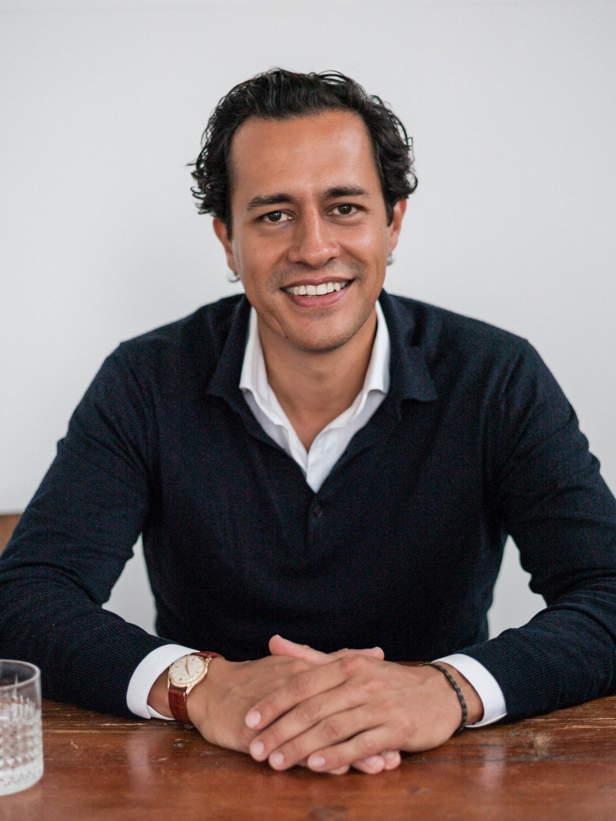 A smiling man with short dark hair in a black sweater and white shirt sitting at a wooden table, with a glass of water nearby.