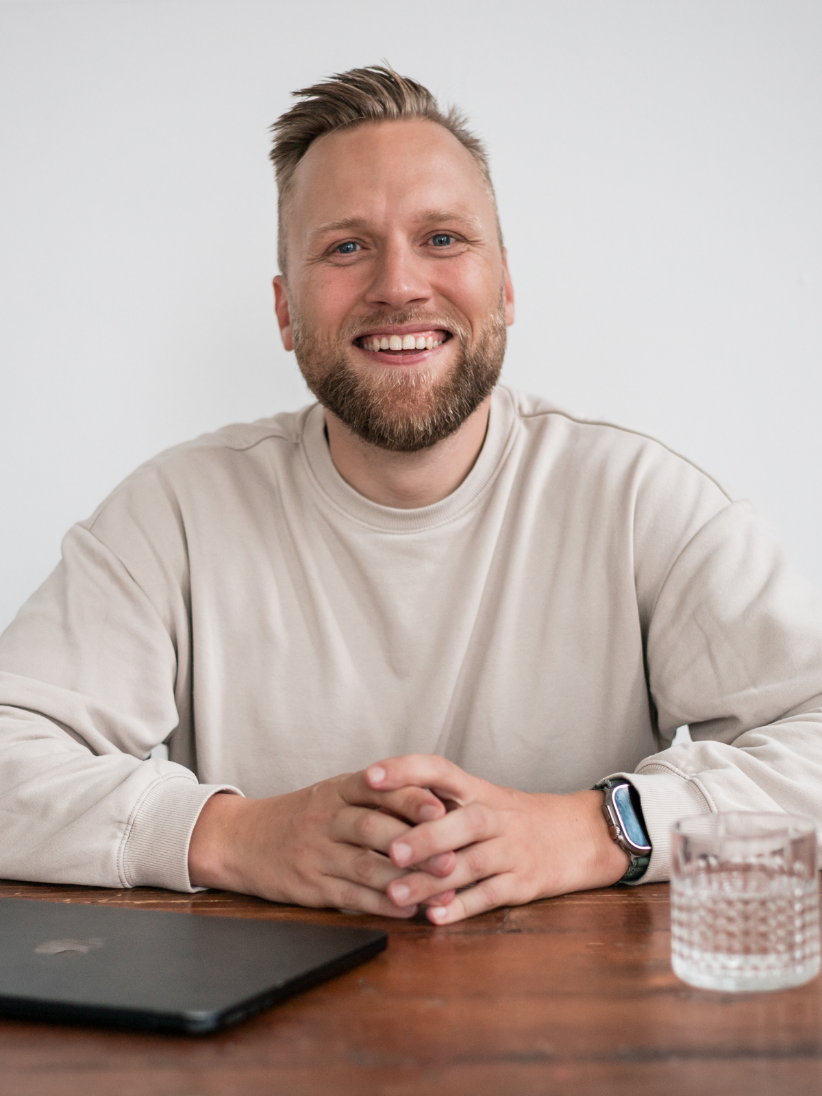 Smiling man with a beard, wearing a beige sweatshirt, sitting at a wooden table with a closed laptop, a glass of water, and a smartwatch.