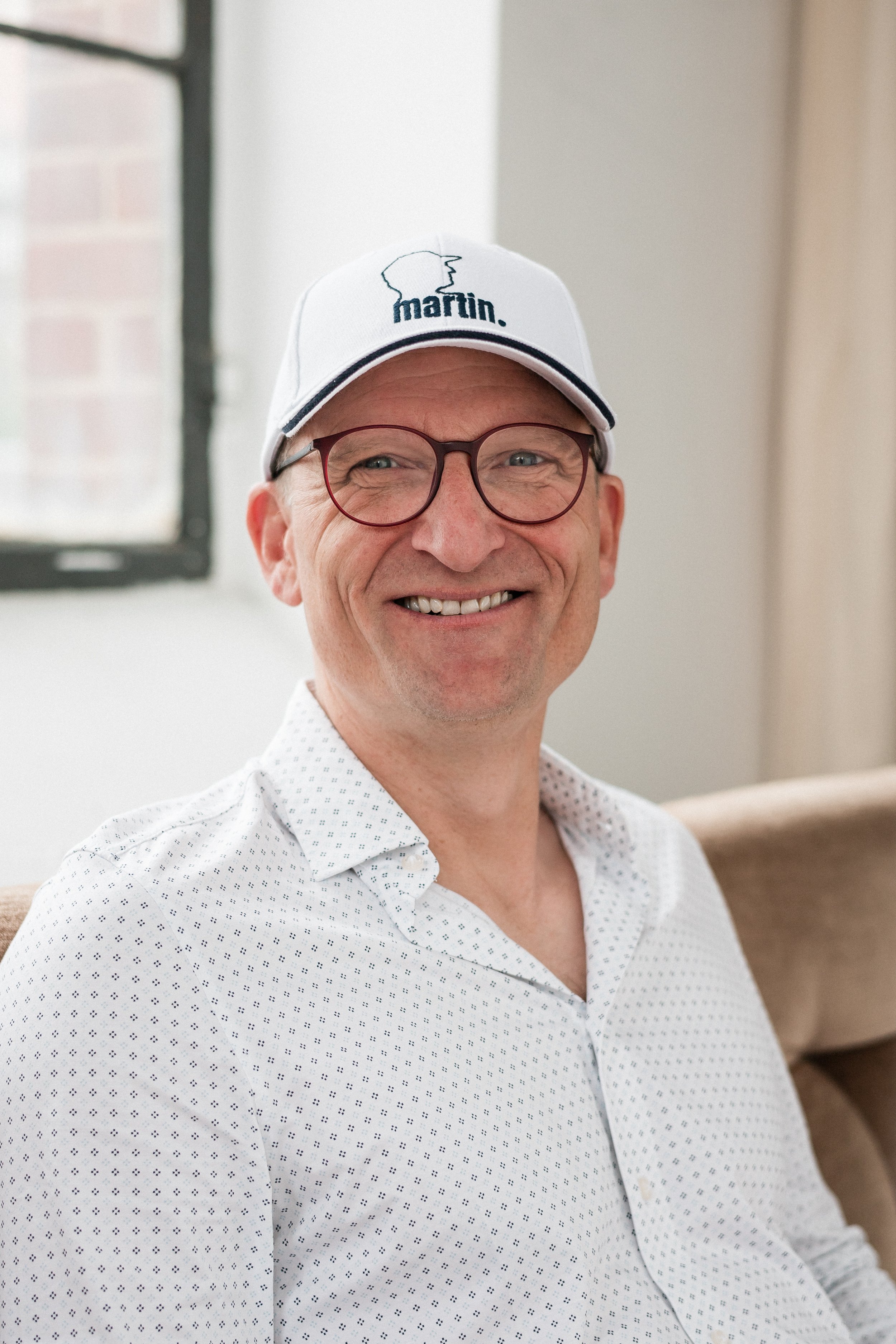 A smiling man wearing glasses, a white patterned shirt, and a white cap with the 'martin.' logo, sitting indoors near a window.