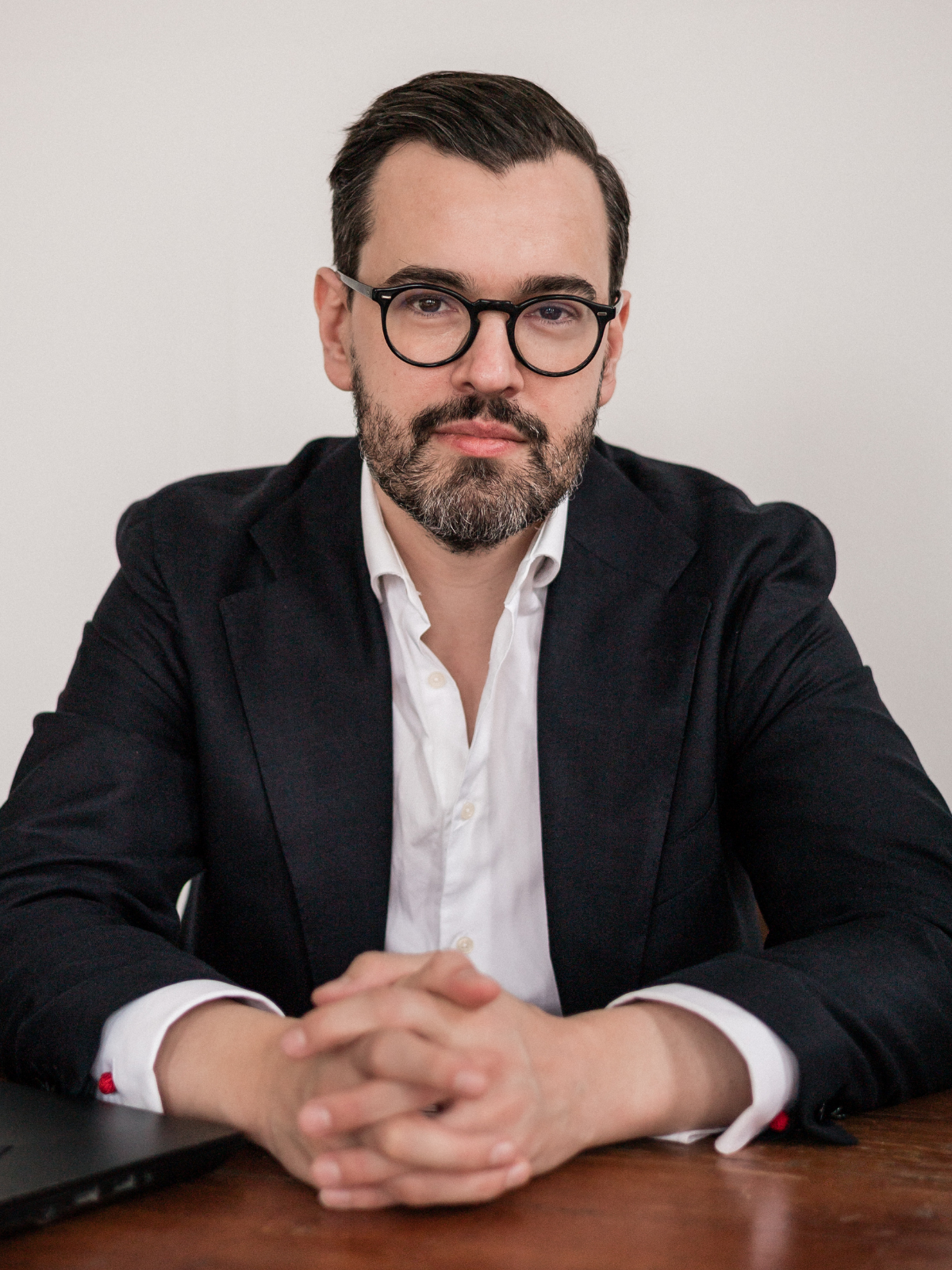 A man with dark hair, glasses, and a beard wearing a white shirt and black blazer, sitting at a wooden table with his hands clasped, in front of a plain white wall.