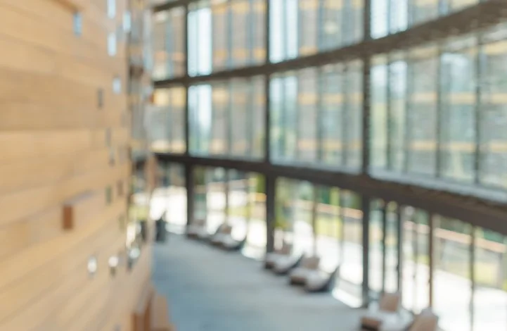 Blurred view of a modern indoor space with large glass windows and a wooden wall, showing some chairs and a corridor.