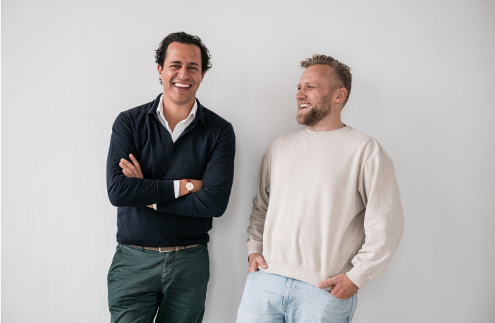 Two men smiling and laughing while standing against a plain white wall.